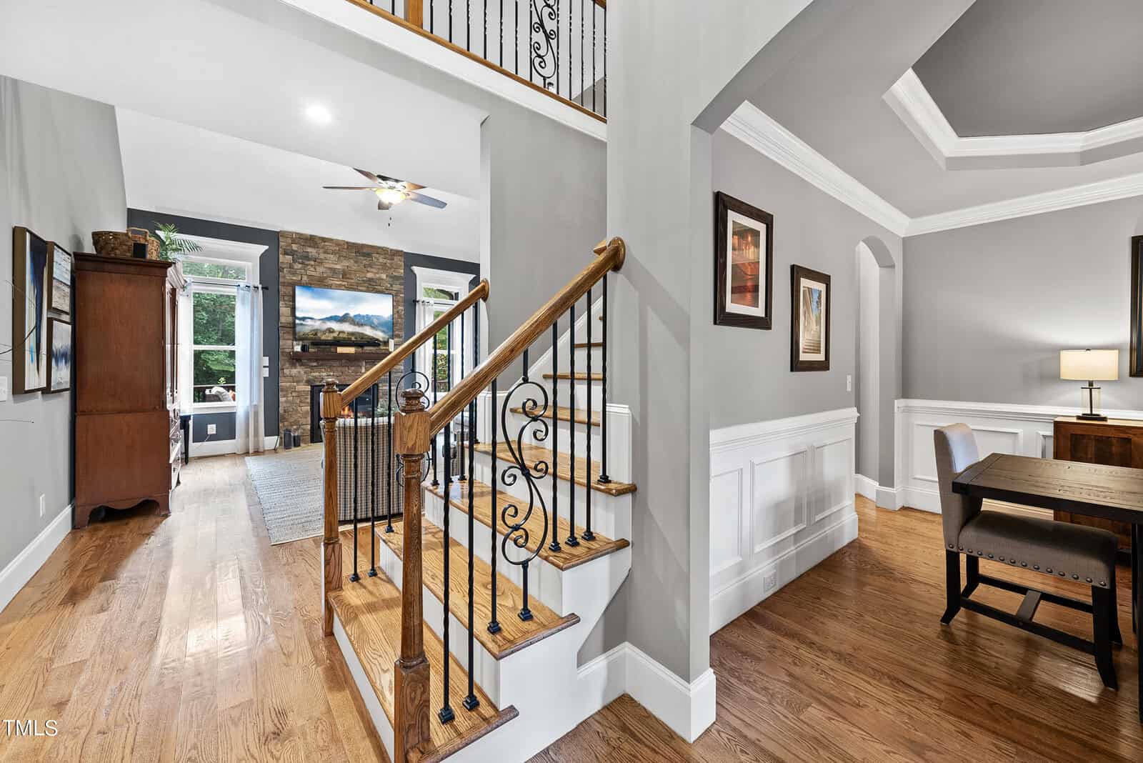 A modern home interior featuring a wooden staircase with ornate iron railings. The open layout includes a living area with a stone fireplace, hardwood floors, and framed artwork on grey walls. A dining table with a lamp is visible on the right side.