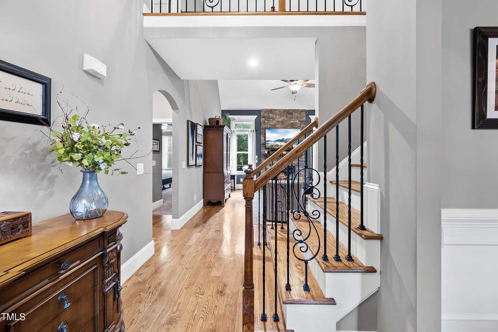 A modern hallway with hardwood floors, a wooden staircase with wrought iron railings, and a vase with flowers on a wooden cabinet. An archway leads to an open living area with a stone fireplace and a ceiling fan in the background.