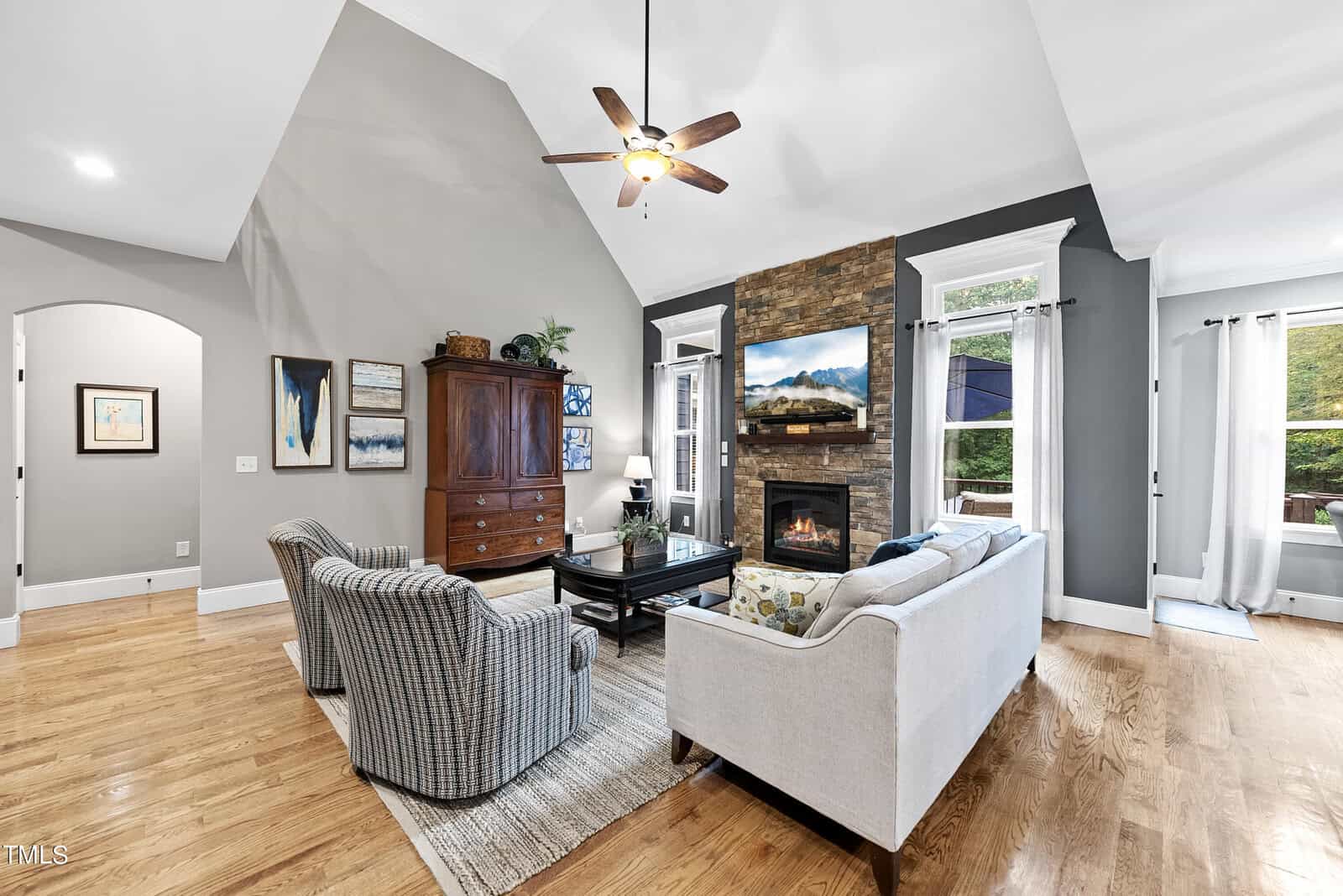 A cozy living room features a high ceiling, ceiling fan, and hardwood floors. There's a stone fireplace, a wooden cabinet, two striped armchairs, and a white sofa with a coffee table and rug. Large windows let in ample natural light.