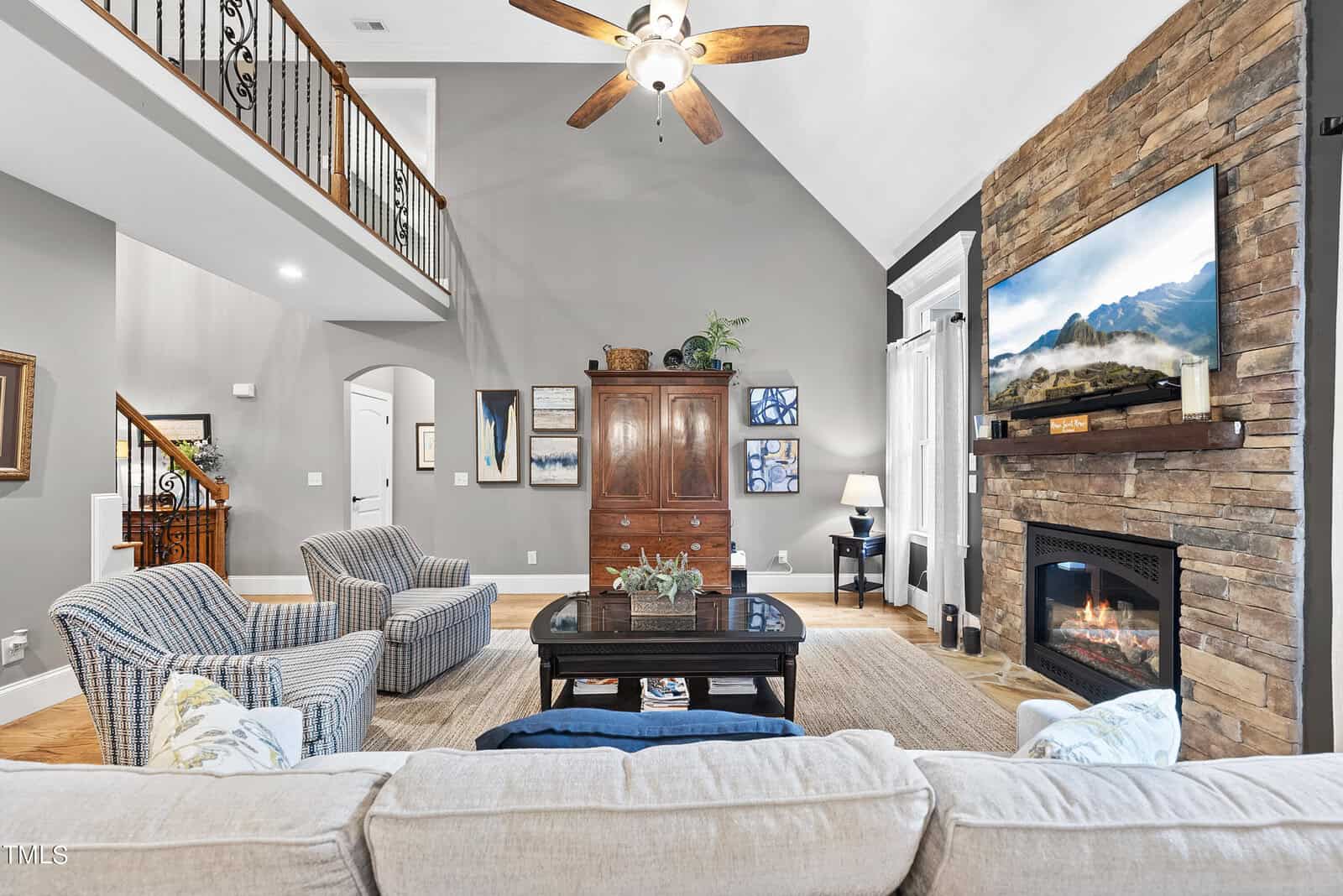 A cozy living room features a stone fireplace with a TV mounted above it, surrounded by beige walls. Two striped armchairs and a beige sofa face a wooden coffee table. A large wooden cabinet adorns the center wall, and a ceiling fan hangs overhead.