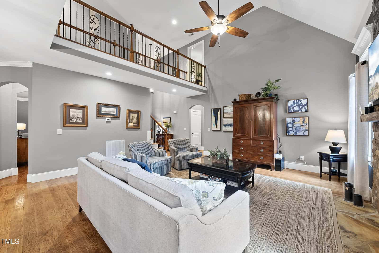 Spacious living room with light gray walls, wooden flooring, and a high ceiling featuring a fan. Includes a white sofa, two blue armchairs, a wooden cabinet, and decorative wall art. Natural light streams through large windows with curtains.