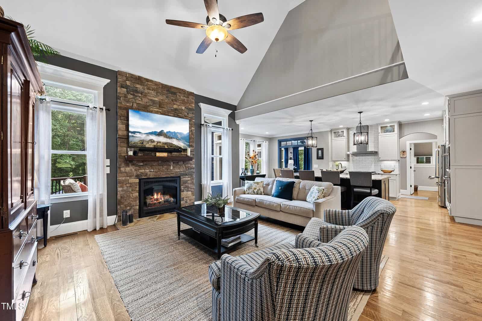 A spacious living room with a vaulted ceiling, featuring a stone fireplace with a TV above, a gray sofa, patterned armchairs, and a coffee table on a woven rug. The room opens to a modern kitchen with an island and bar stools. Large windows bring in natural light.