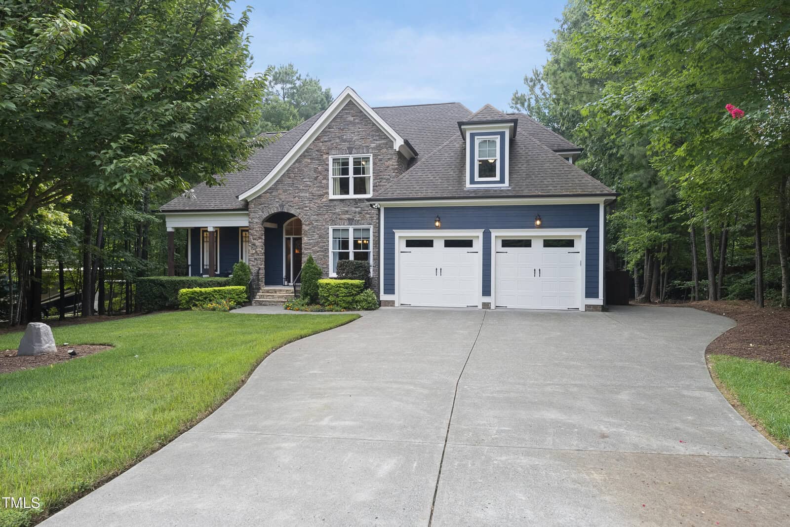 A modern home with stone and blue siding, featuring a gabled roof and large white double garage doors. The house is surrounded by lush greenery and a well-kept lawn, with a wide driveway leading to the entrance.