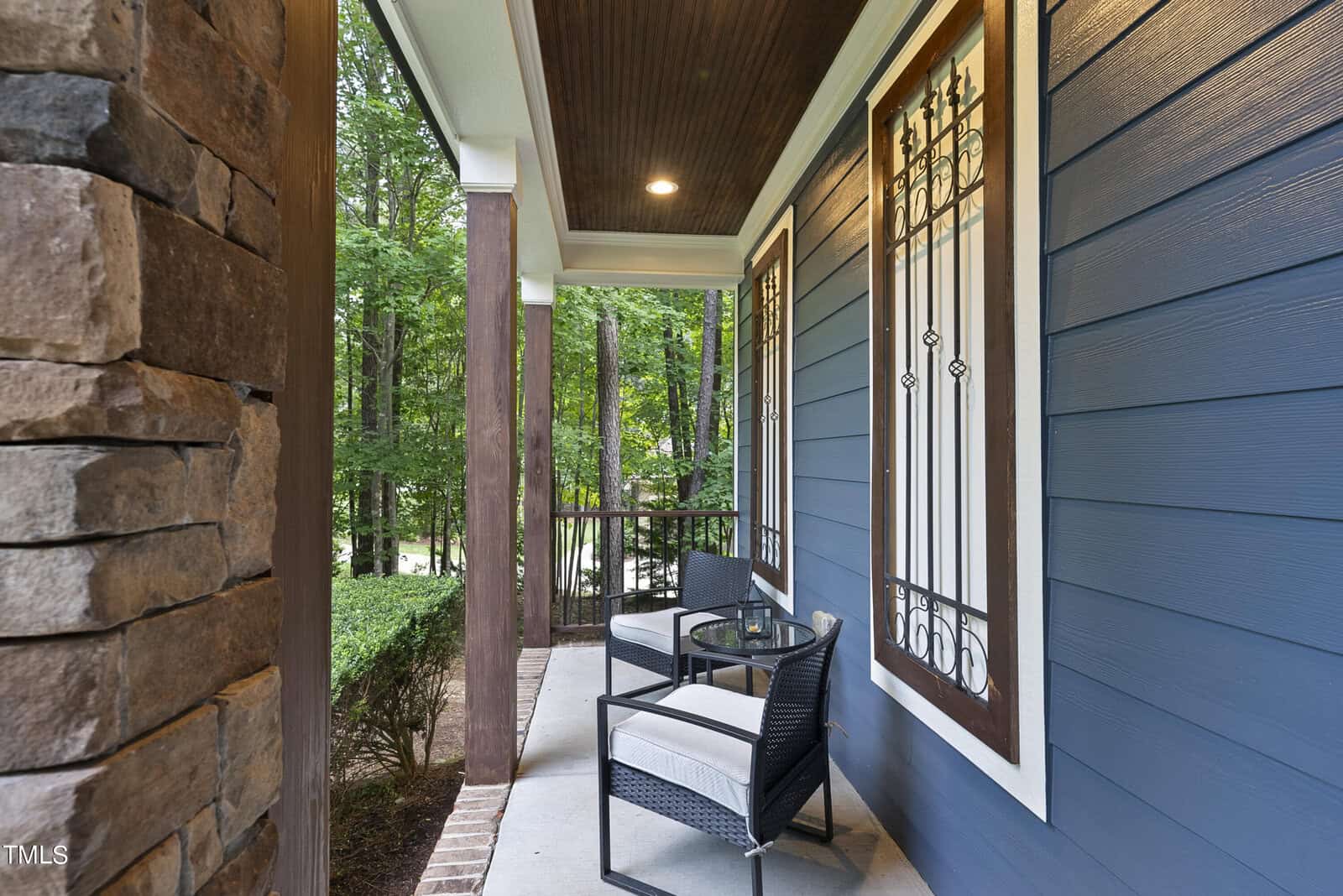 A cozy porch with two cushioned chairs and a round glass table. The porch is framed by wooden pillars and stone accents, with a view of lush green trees in the background. A dark wooden ceiling and deep blue siding complete the aesthetic.