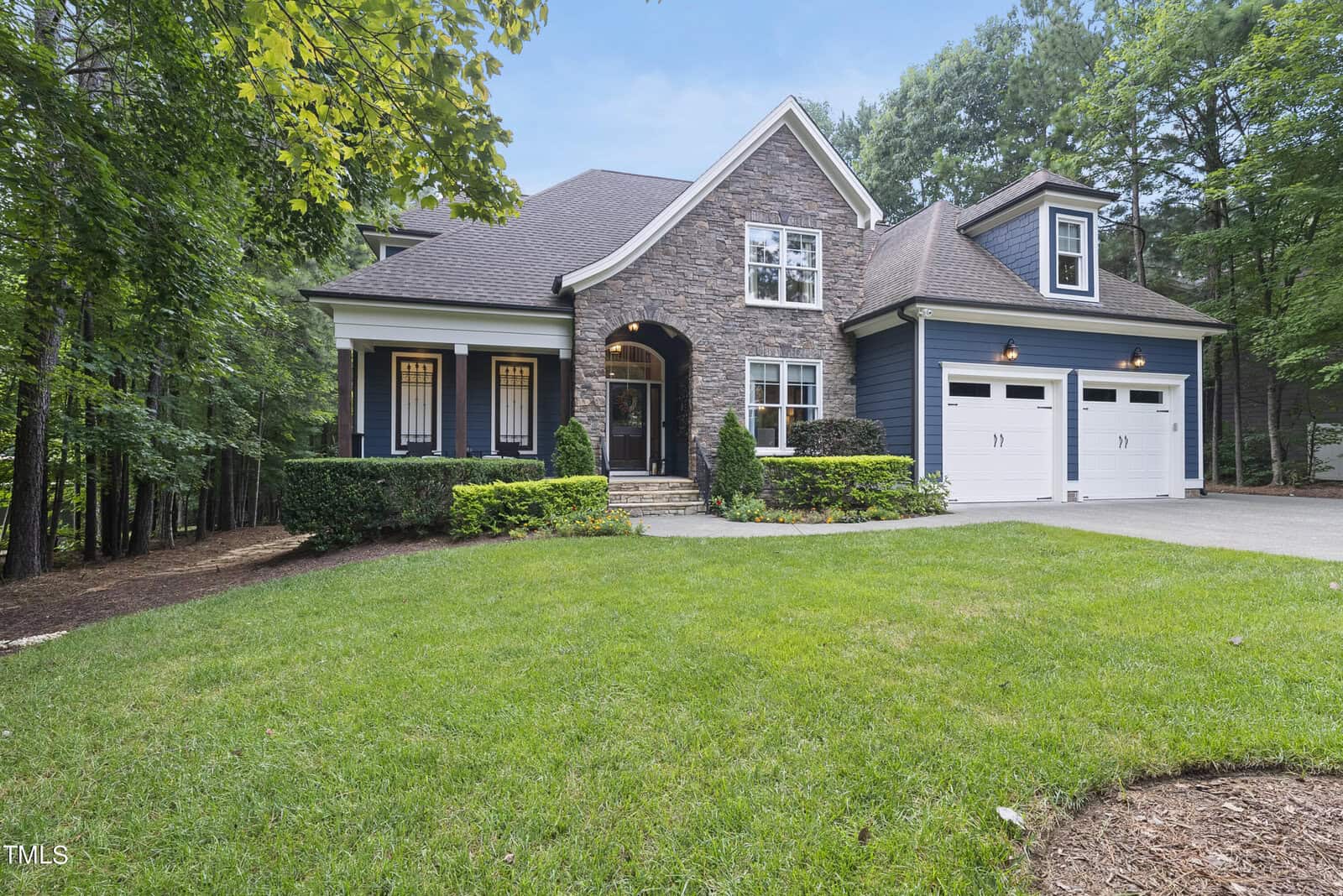 Nestled in Wake Forest at 508 Belmont Circle, this charming two-story house boasts a stone facade and blue siding, surrounded by lush greenery. It features a spacious grassy lawn, double garage, and paved driveway under the clear blue sky.