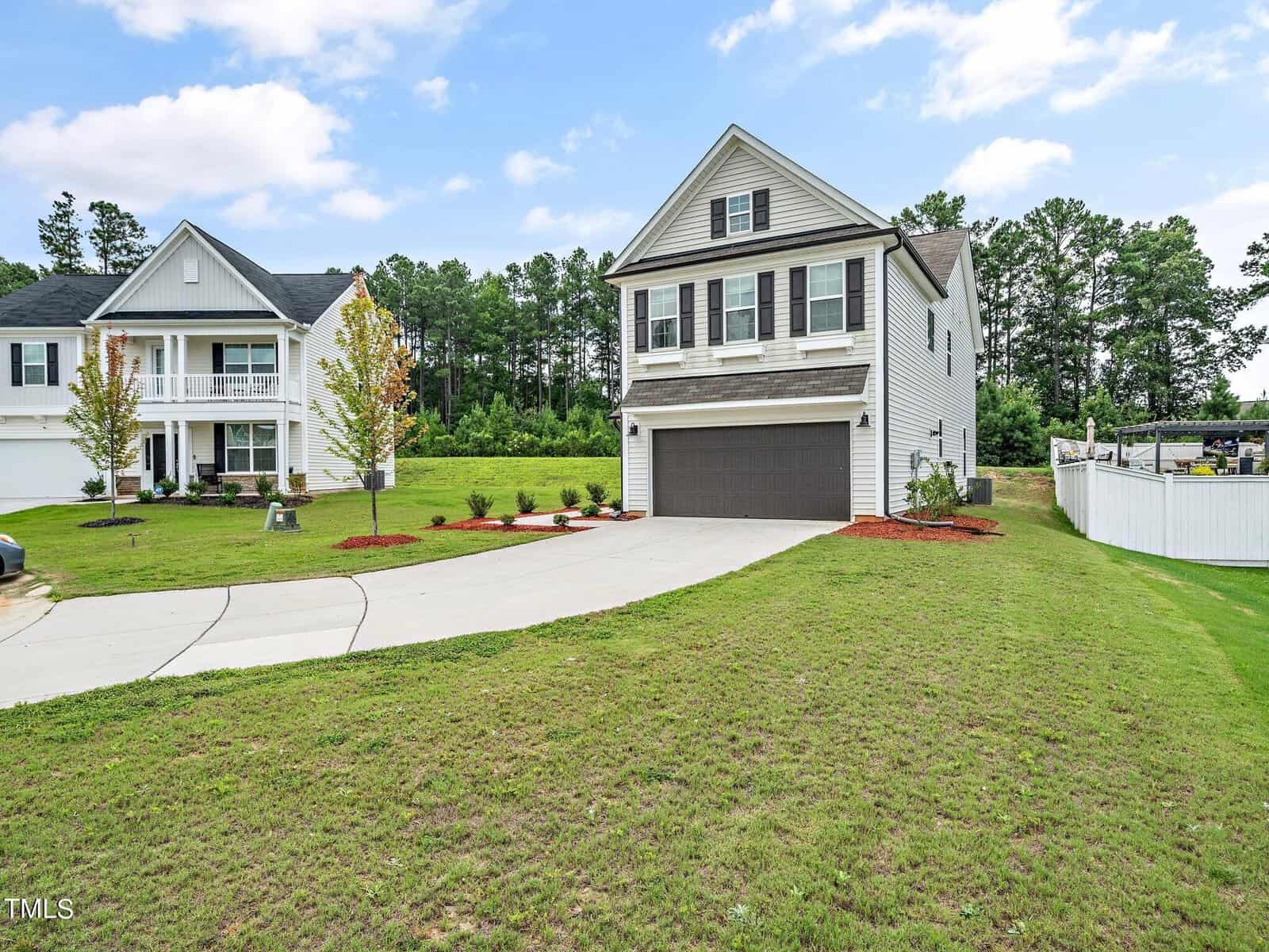A two-story suburban house with gray siding, a double garage, and a well-kept lawn, sits next to a similar house. The yard features young trees, shrubs, and a white fence with woods in the background under a partly cloudy sky.