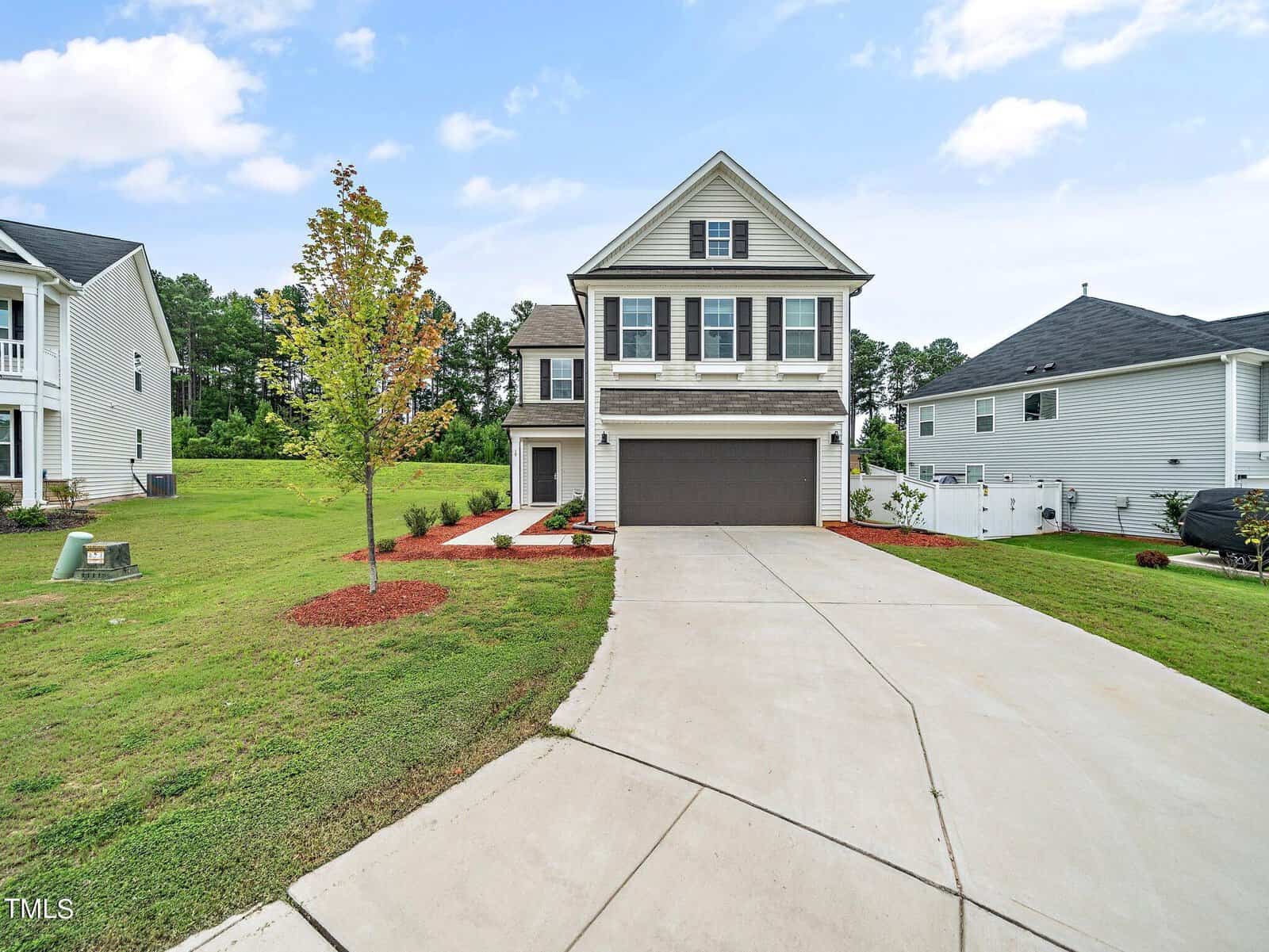 A modern two-story house with gray siding, black shutters, and a double garage sits at the end of a curved driveway. The front yard has a small tree, red mulch, and green grass, with neighboring houses nearby.