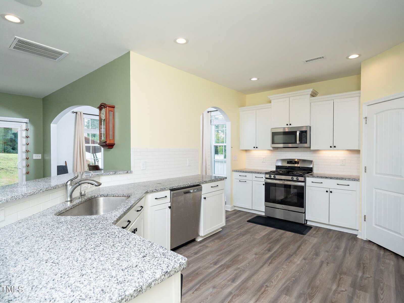 Modern kitchen with white cabinets, stainless steel appliances, granite countertops, light yellow and green walls, subway tile backsplash, and wood-look flooring; open layout with large windows and arched doorways.