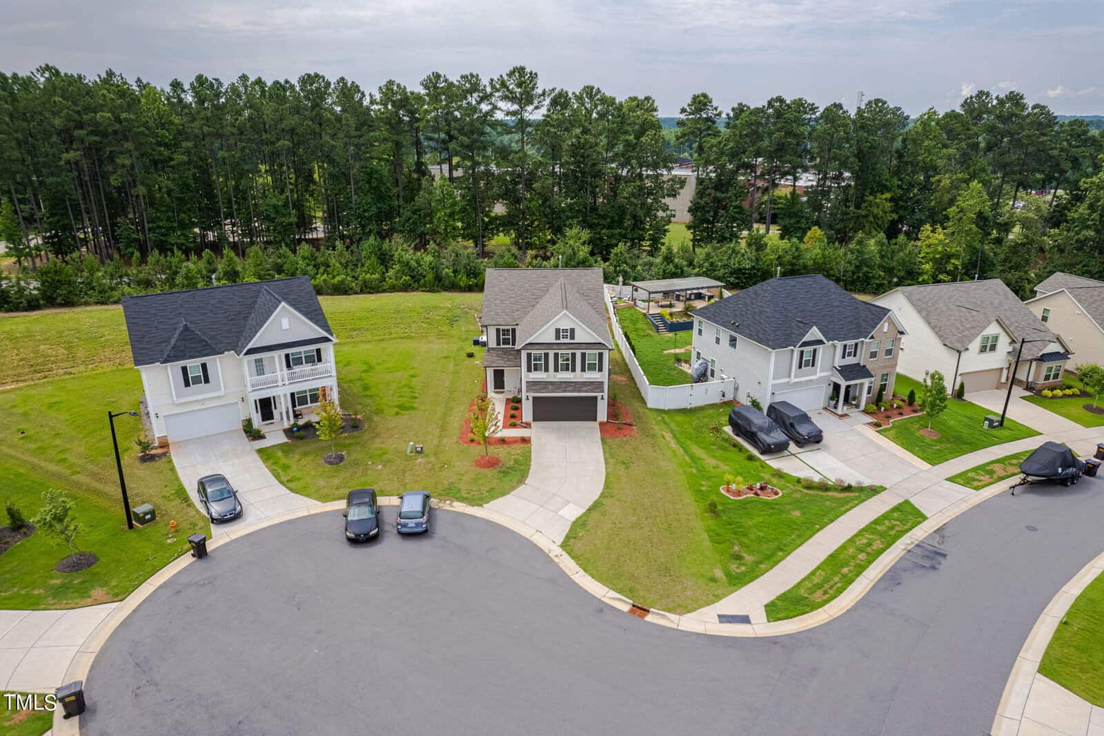 Aerial view of a suburban cul-de-sac with five modern two-story houses, three driveways with cars parked, well-maintained lawns, and a backdrop of tall green trees.