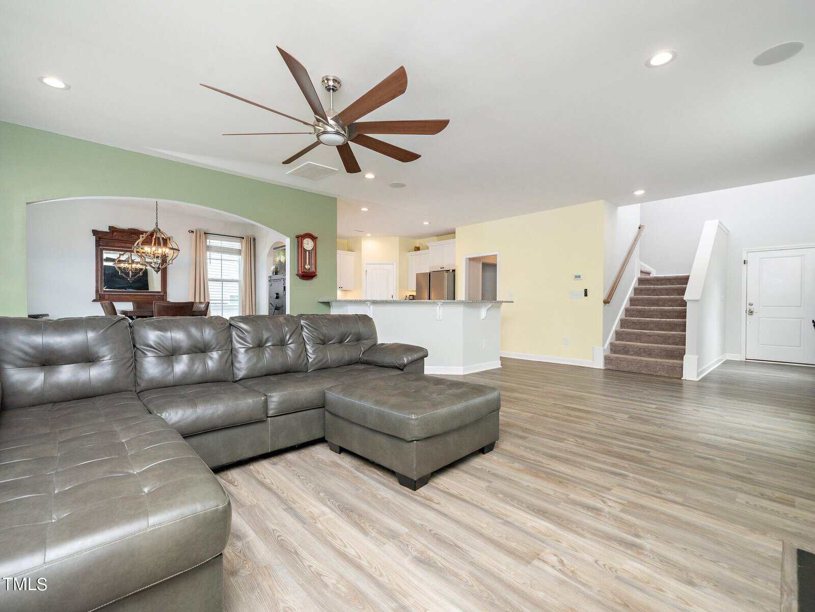 Spacious living room with a large gray sectional sofa and ottoman, wood flooring, ceiling fan, open kitchen with white cabinets, dining area, green accent wall, and staircase leading to the upper floor.
