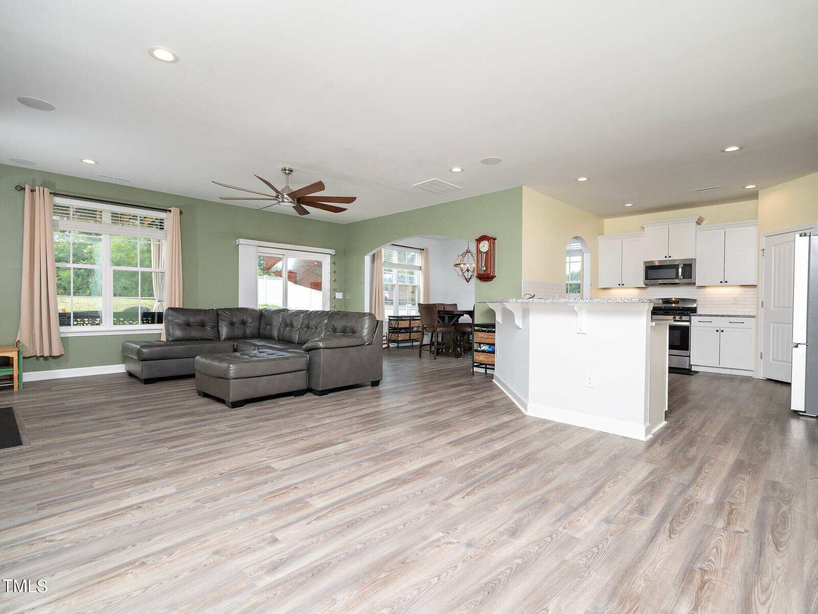 Open-concept living area with gray sectional sofa, large windows, ceiling fan, and light wood flooring; adjacent to a modern kitchen with white cabinets, stainless steel appliances, and a breakfast bar.