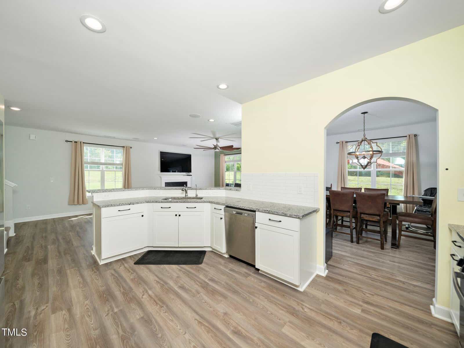 Modern kitchen with white cabinets, granite countertops, and stainless steel dishwasher. Open layout connects to living room with fireplace and dining room visible through an arched doorway. Wood-look flooring throughout.