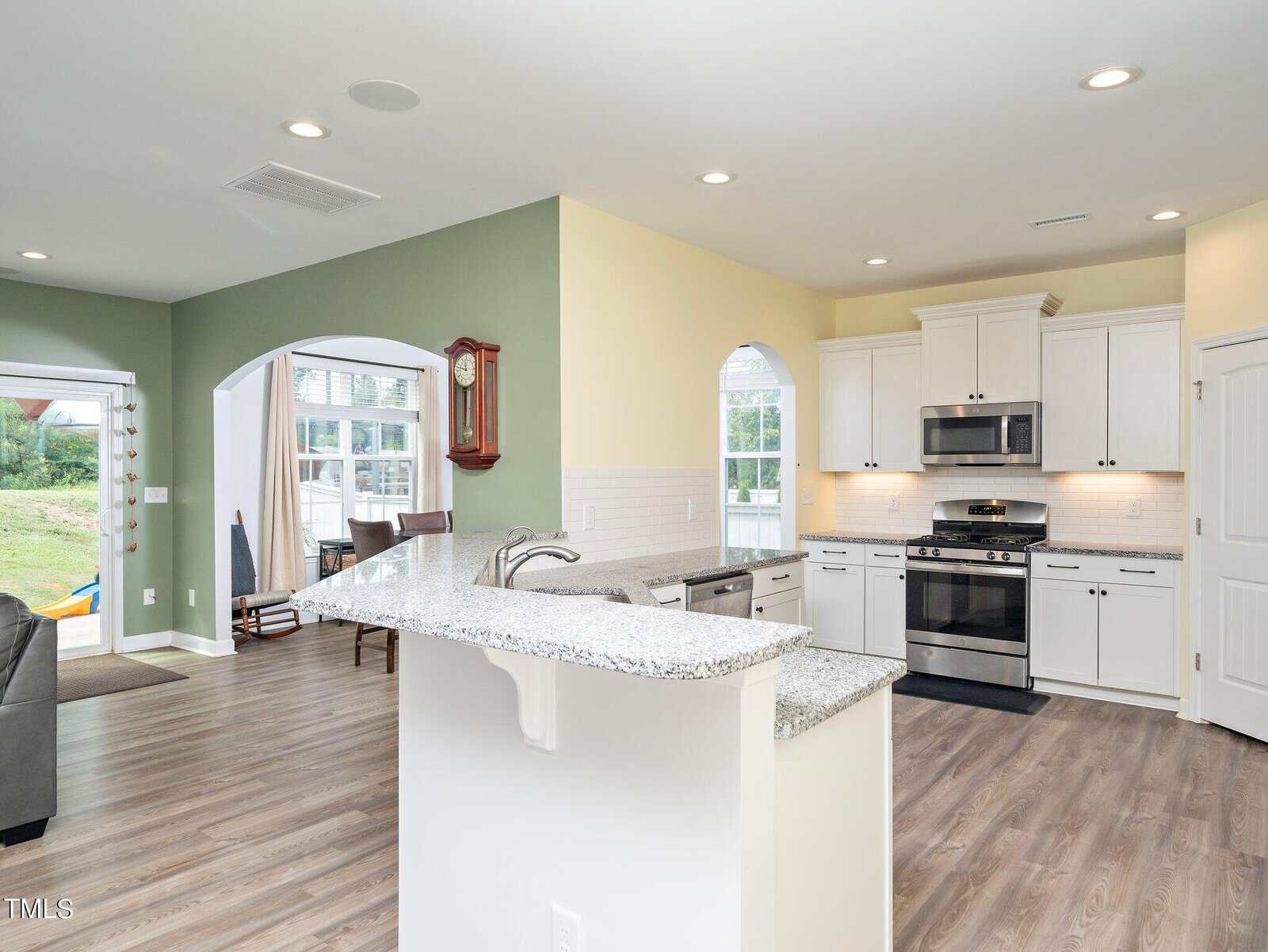 Modern open-concept kitchen with white cabinets, stainless steel appliances, granite countertops, and wood flooring. Kitchen opens to a living area with green accent wall, clock, and large windows with views of the yard.