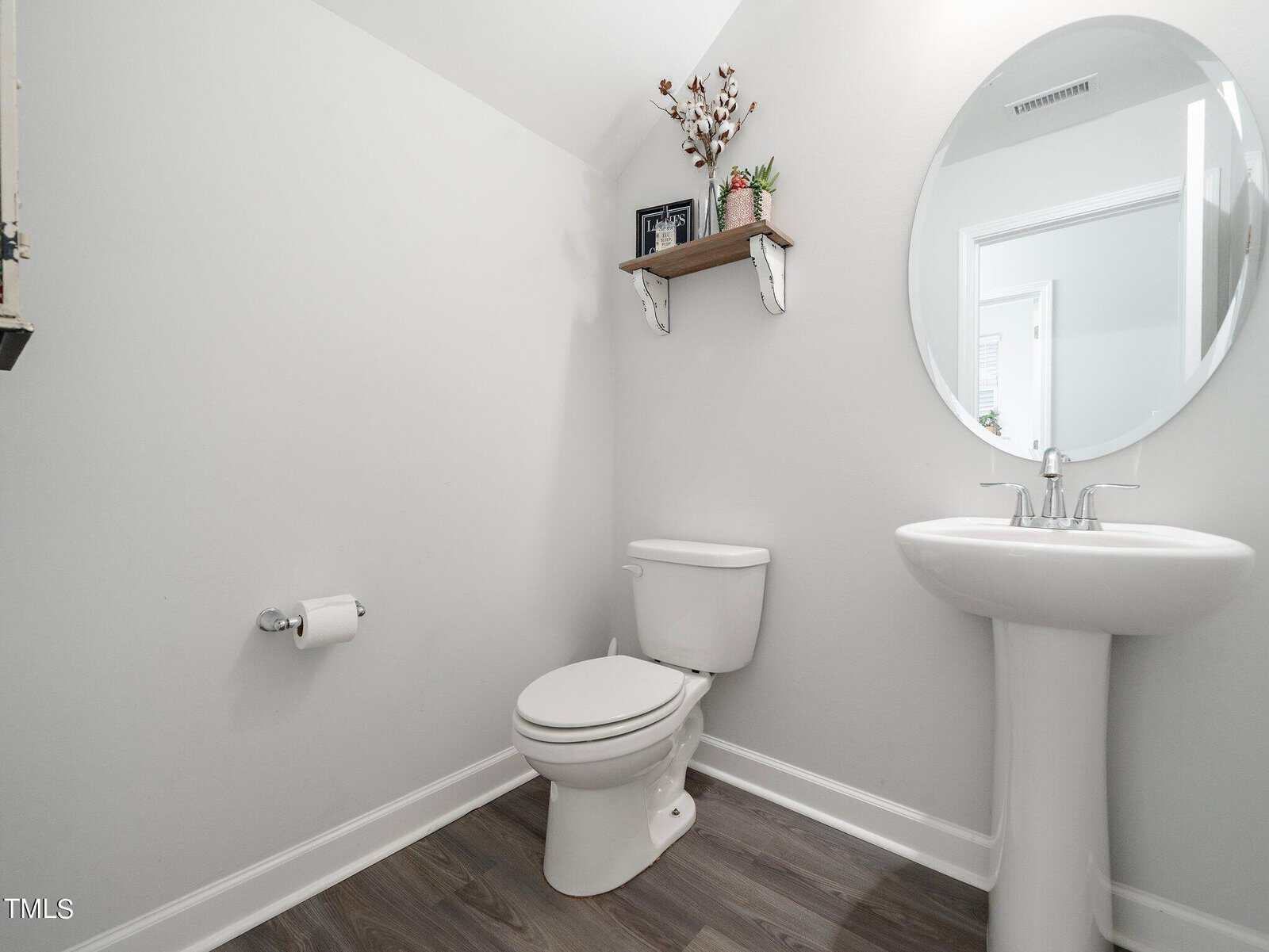 A small bathroom with white walls, a pedestal sink, a toilet, a round mirror, a toilet paper holder, and a wall shelf holding decorative items and small plants. The floor has a dark wood finish.