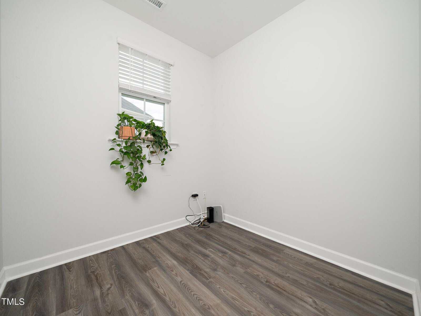 Empty white room with gray wood flooring, a small window with white blinds, a potted plant on the windowsill, and some electronic devices with cords on the floor in the corner.