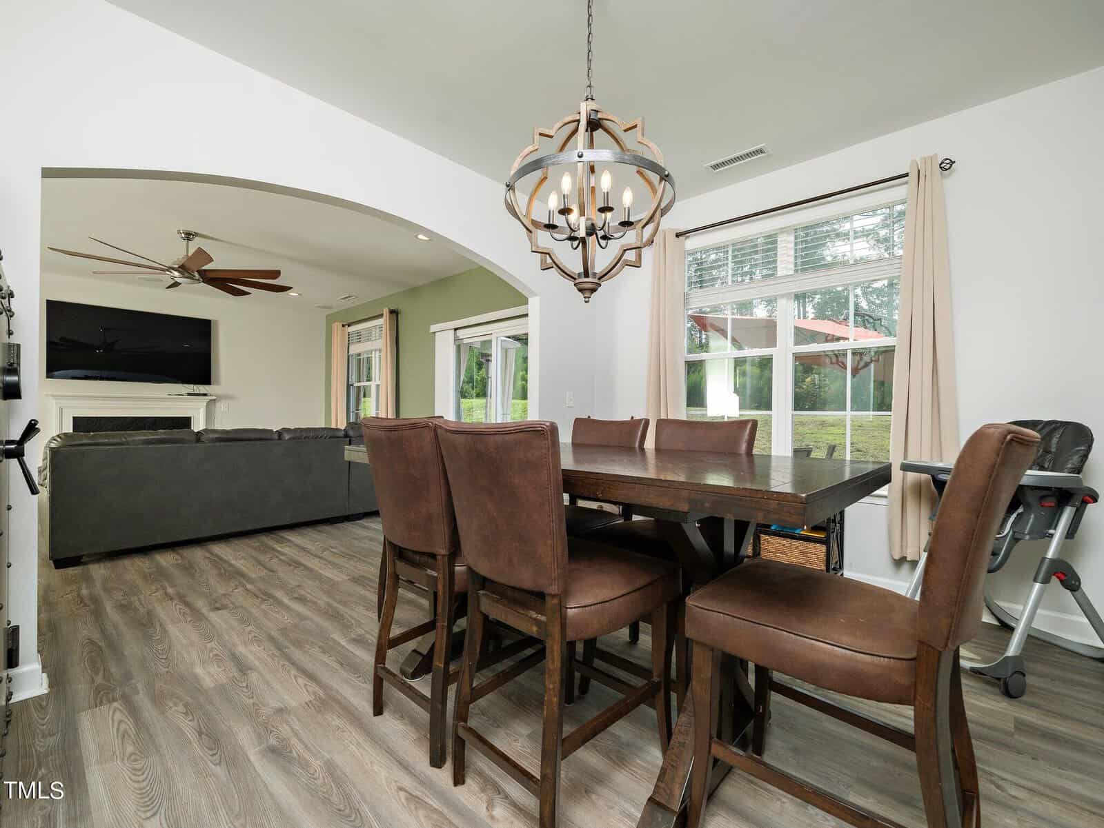 A modern dining area with a dark wooden table and six brown leather chairs, adjacent to a living room with a gray sofa, ceiling fan, and fireplace. Large windows provide views of a green yard and outdoor umbrella.