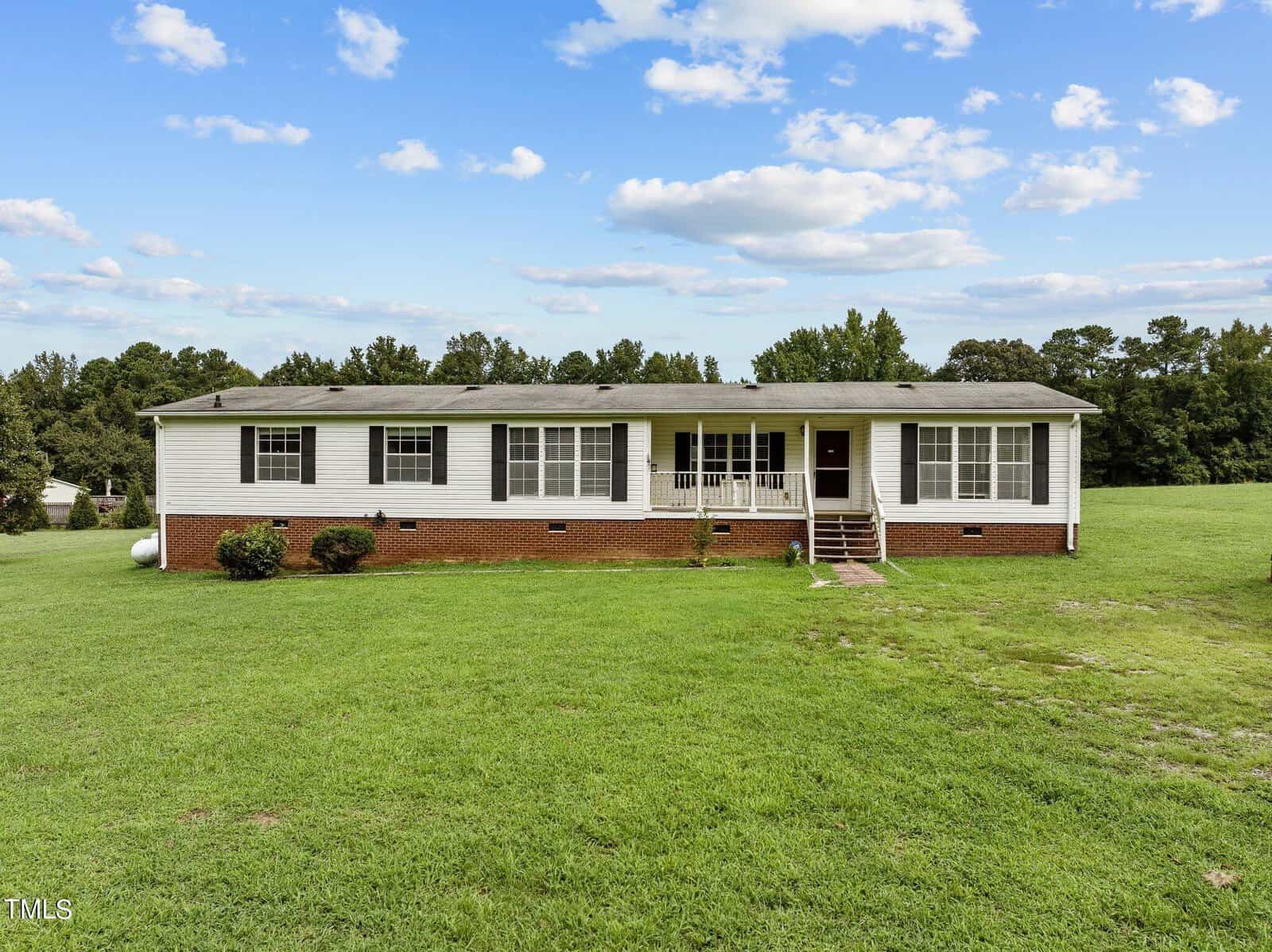 A single-story house with white siding and a red brick foundation sits on a large, well-maintained lawn. The home features multiple windows, a front porch with white railings, and steps leading up to the entrance. The sky is partly cloudy with a few scattered clouds.