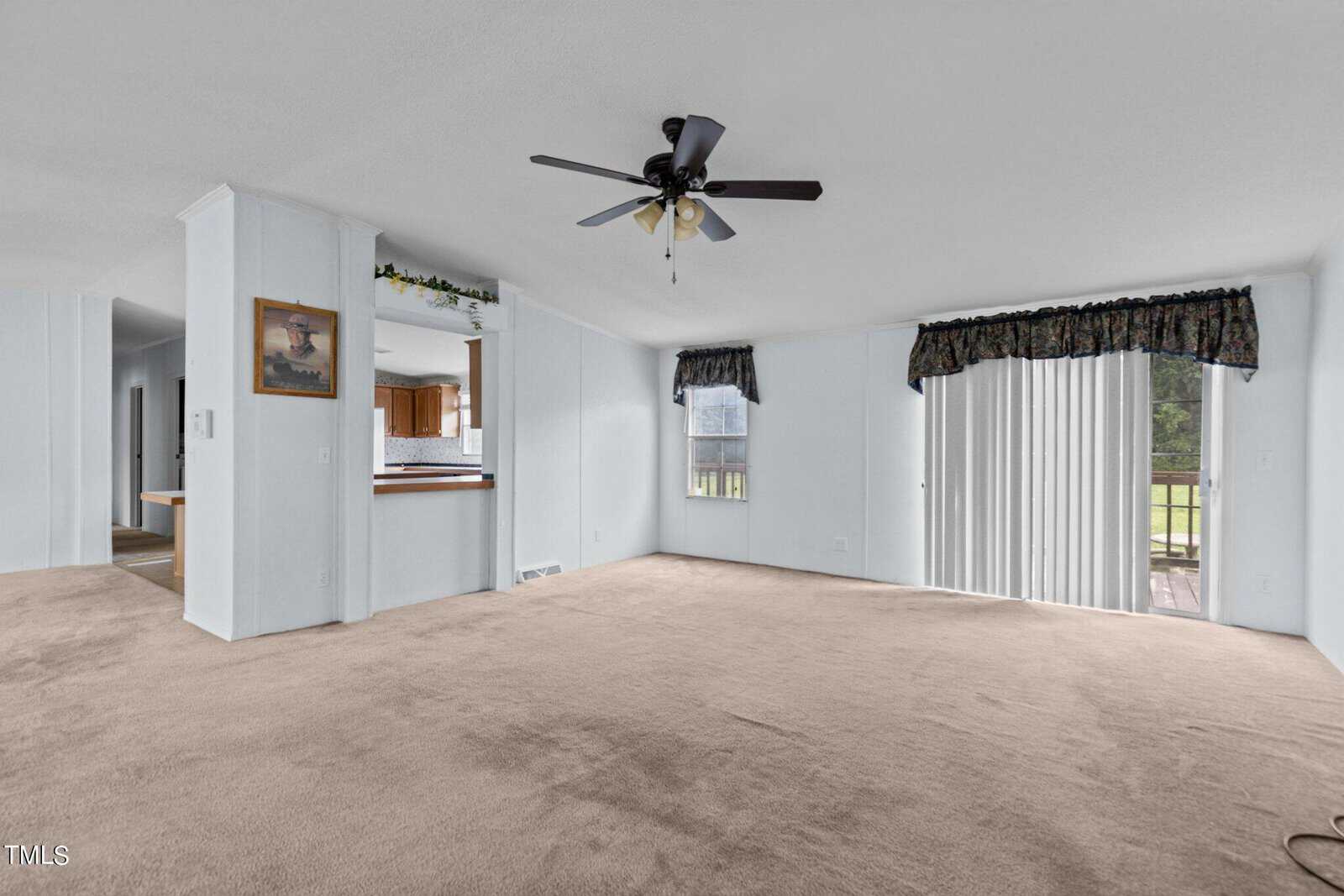 A spacious living room with light blue walls and beige carpet. It features a ceiling fan with light in the middle, an open view to the kitchen area, a framed painting on the wall, and large windows with dark floral valances, including sliding doors leading outside.