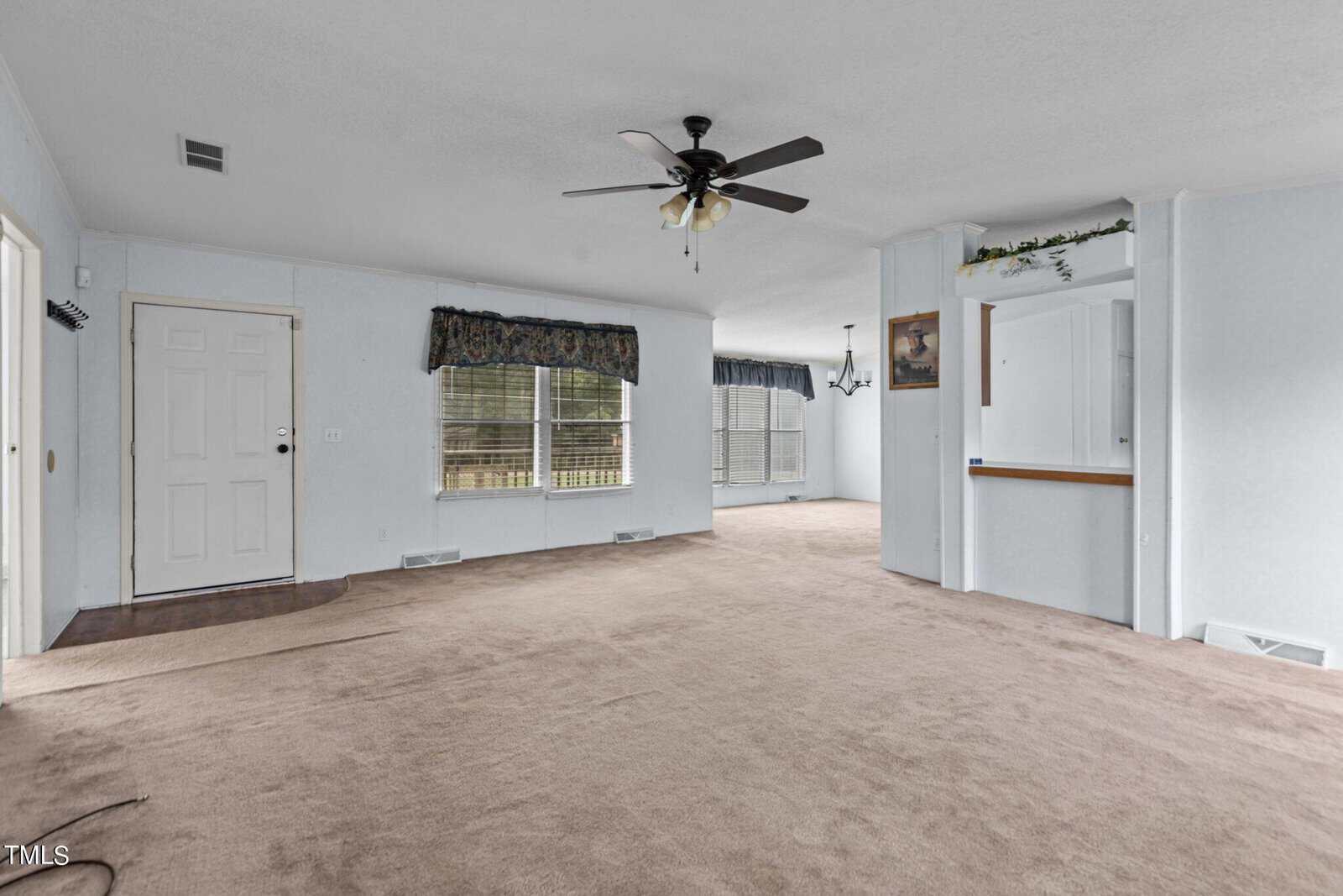 A spacious, empty living room with light blue walls and beige carpet. A ceiling fan with light fixtures hangs from the white ceiling. Large windows with patterned valances provide natural light. An open doorway leads to another room with additional windows and a chandelier.