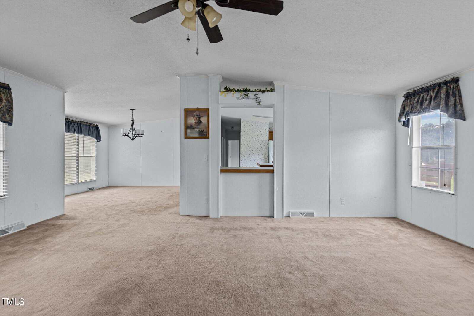 Spacious living room with light-colored walls and carpet. Ceiling fan with light fixture in the foreground. Connected dining area with chandelier and large windows. Partition wall with a pass-through opening to the kitchen. Blue curtains on windows.