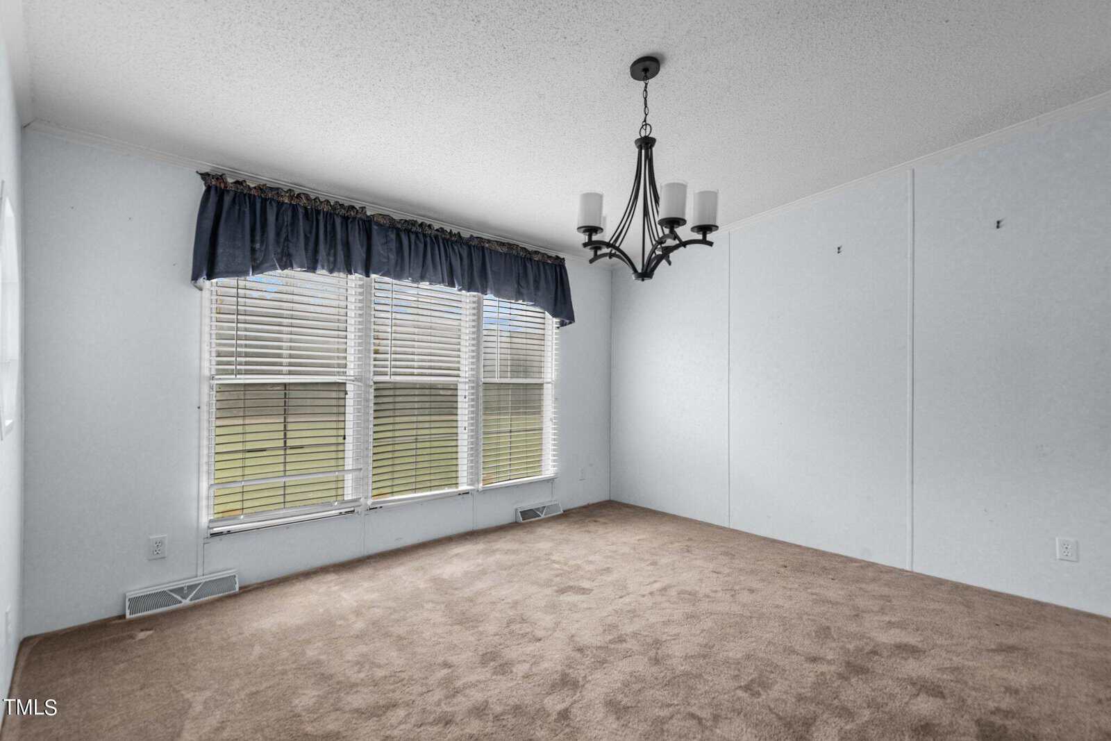 An empty room with pale blue walls, a brown carpeted floor, and a large window with closed blinds and a dark valance. A black metal chandelier with candles hangs from the ceiling. Light is filtering through the window, giving the room a bright appearance.