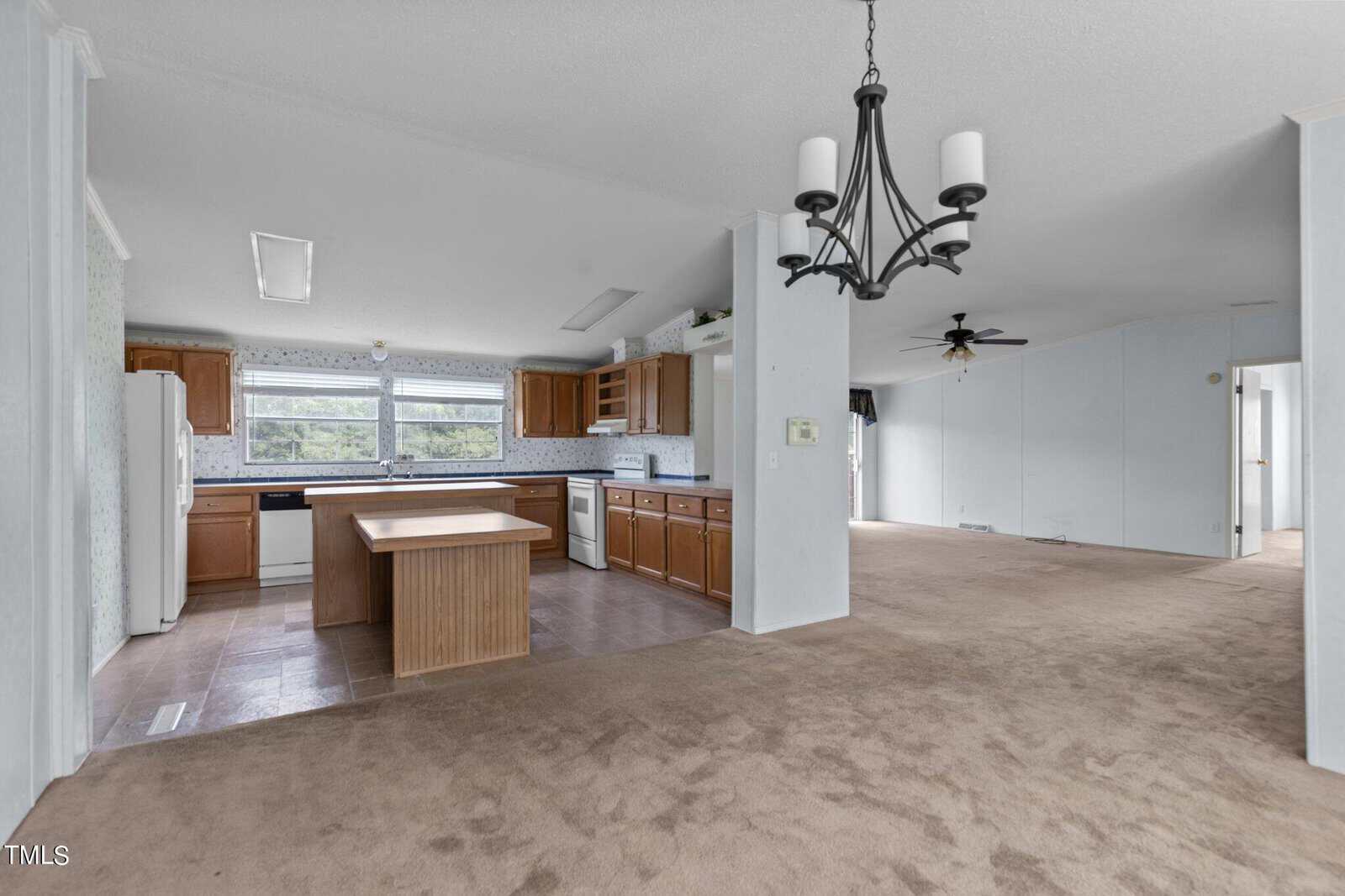 A spacious open-plan kitchen and living area with brown cabinets, white appliances, and an island counter in the kitchen. The living area features beige carpeting, a black ceiling fan, and a black chandelier. Large windows provide ample natural light.