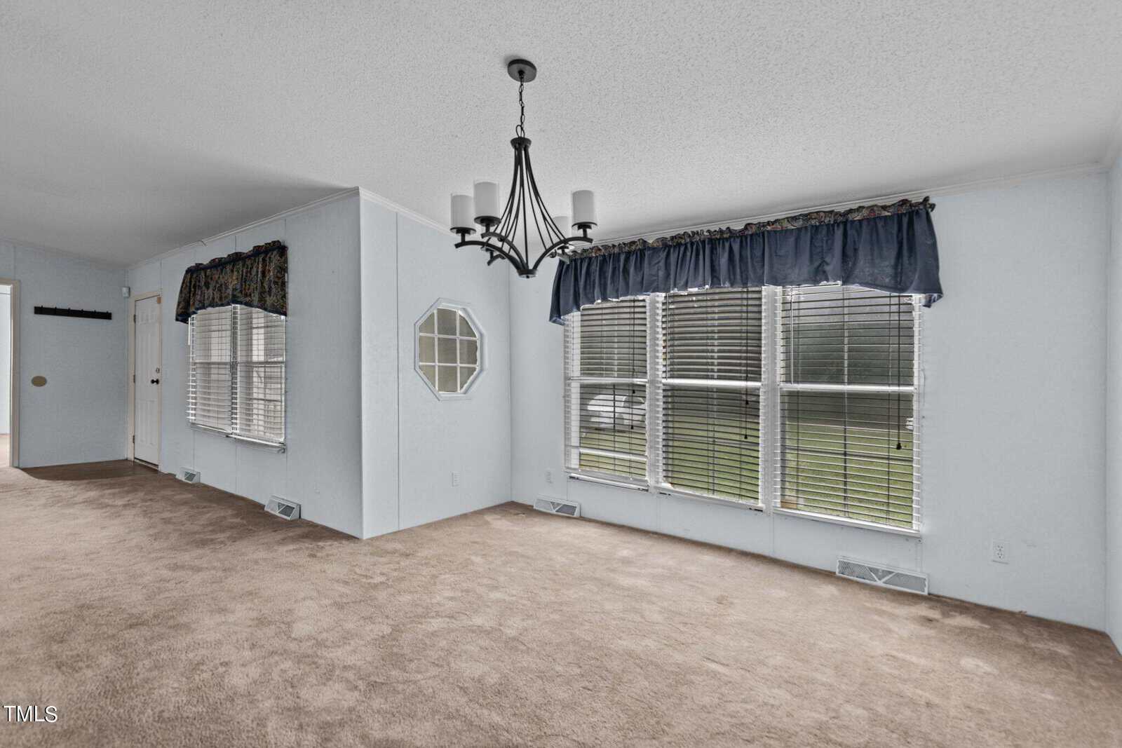 A sunlit living room with light blue walls, beige carpet flooring, and two large windows covered by blue valances. A black chandelier hangs from the ceiling. A small octagonal window is on the wall near the door, which leads to another room.