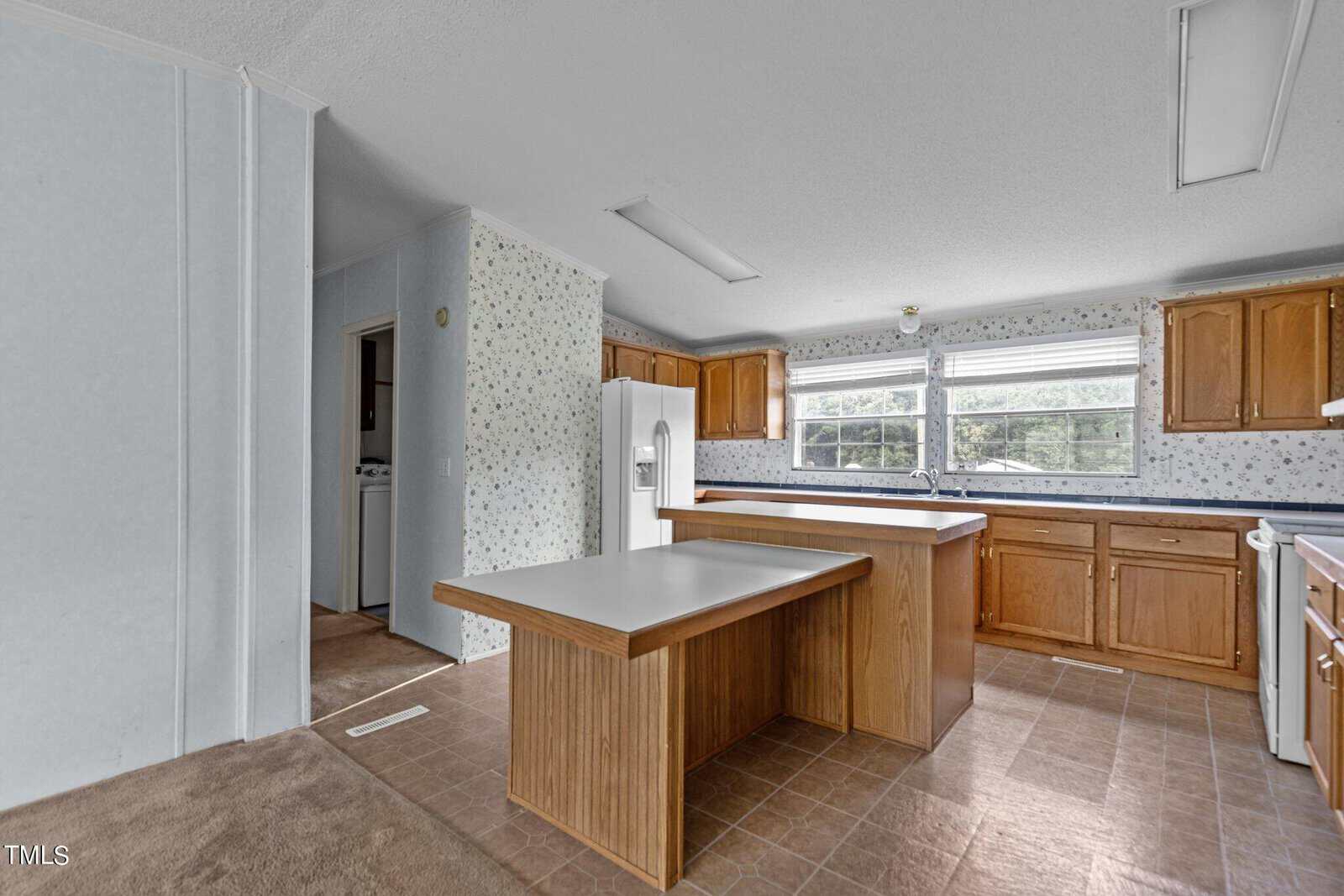 A kitchen with light brown cabinets, white appliances, and a center island. The walls feature floral wallpaper, and large windows bring in natural light. A doorway to the left leads to a laundry area. The room is well-lit with overhead fluorescent lights.