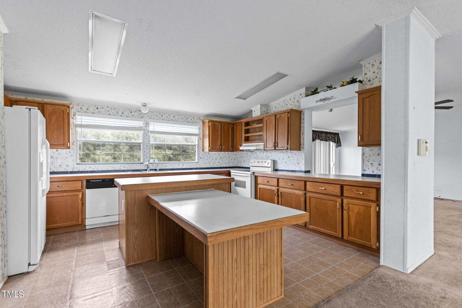 A spacious kitchen with wooden cabinets, white appliances, and a center island with a countertop. The kitchen has tiled flooring, large windows letting in natural light, and floral wallpaper with a white background. There is also an adjacent view of a carpeted area.