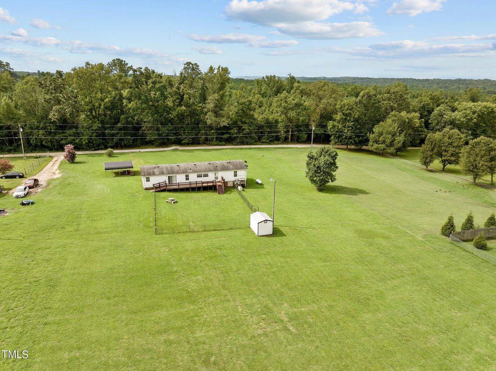 Aerial view of a large, grassy property at 402 Buffalo Creek Lane, Kittrell, featuring a white single-story house with a fenced backyard. A small shed is located within the fenced area. The property is surrounded by lush trees and a distant forest under a partly cloudy sky.