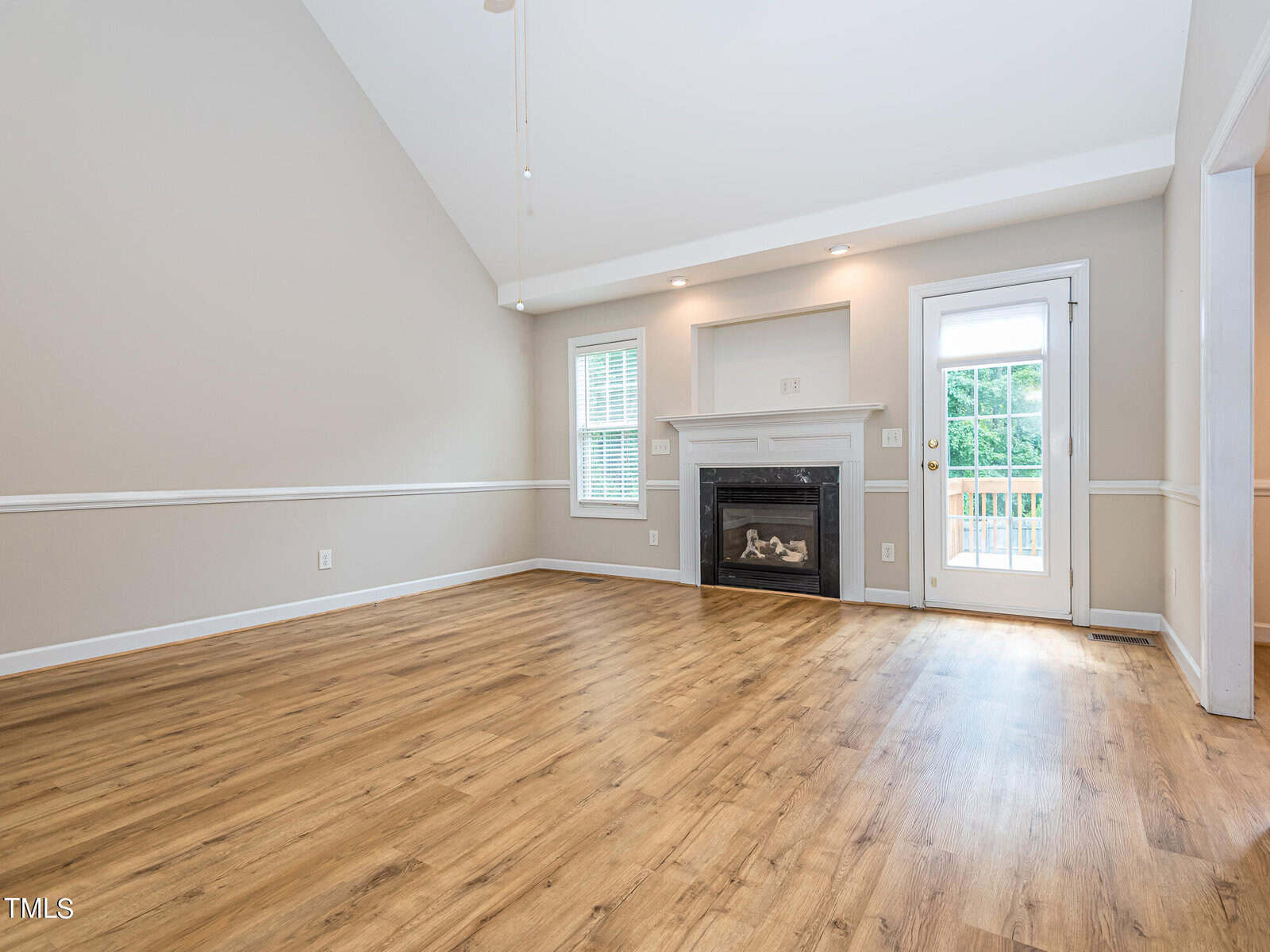 Spacious, empty living room with light wooden flooring and vaulted ceiling. A fireplace is centered on one wall, next to a window. A glass door leads to an outdoor deck, allowing ample natural light. Walls are painted light beige with white trim.