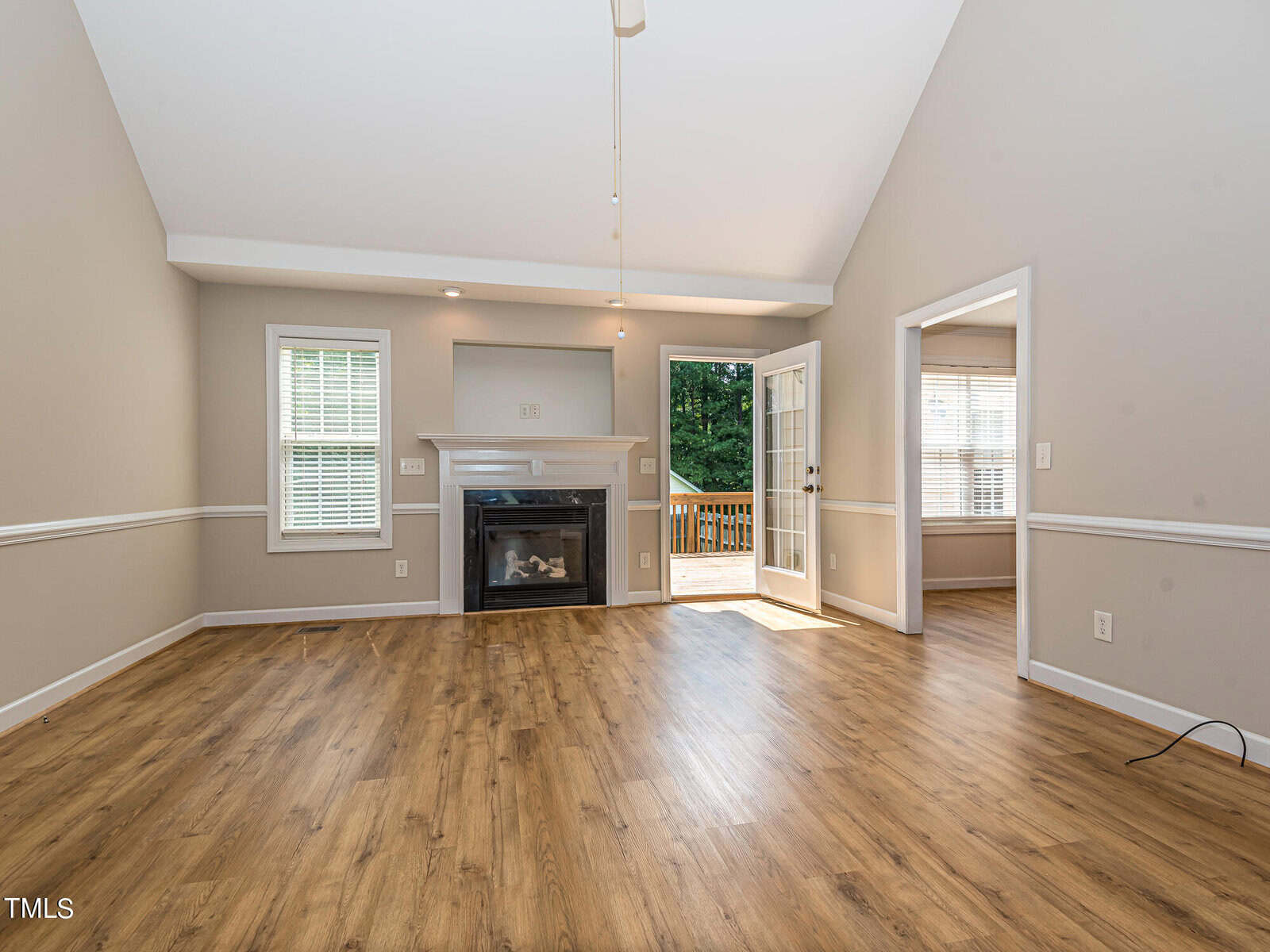 Spacious living room with laminate wood flooring, a fireplace, and two windows. French doors lead to a deck with a view of greenery. Neutral walls and white trim create an airy feel.