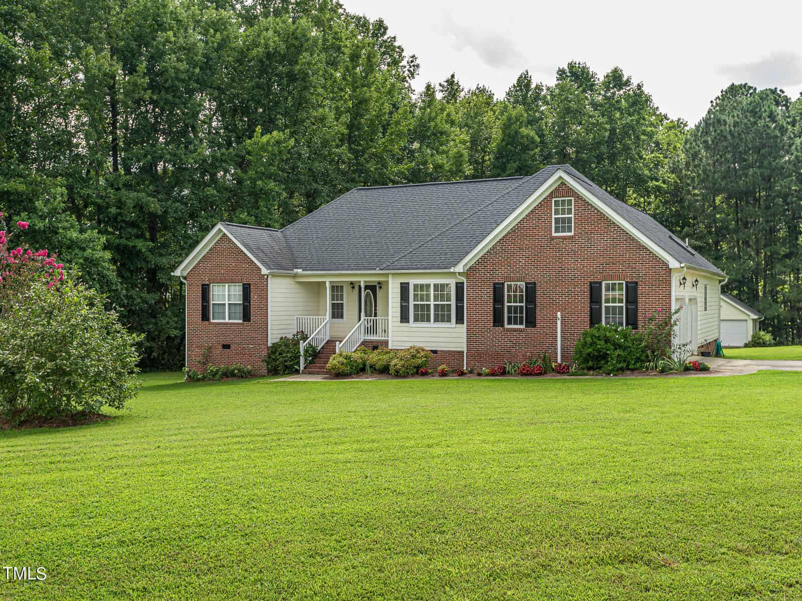 A single-story brick and beige house with a neatly manicured lawn and small garden in front. It has a dark roof and is situated against a backdrop of lush green trees under a cloudy sky.