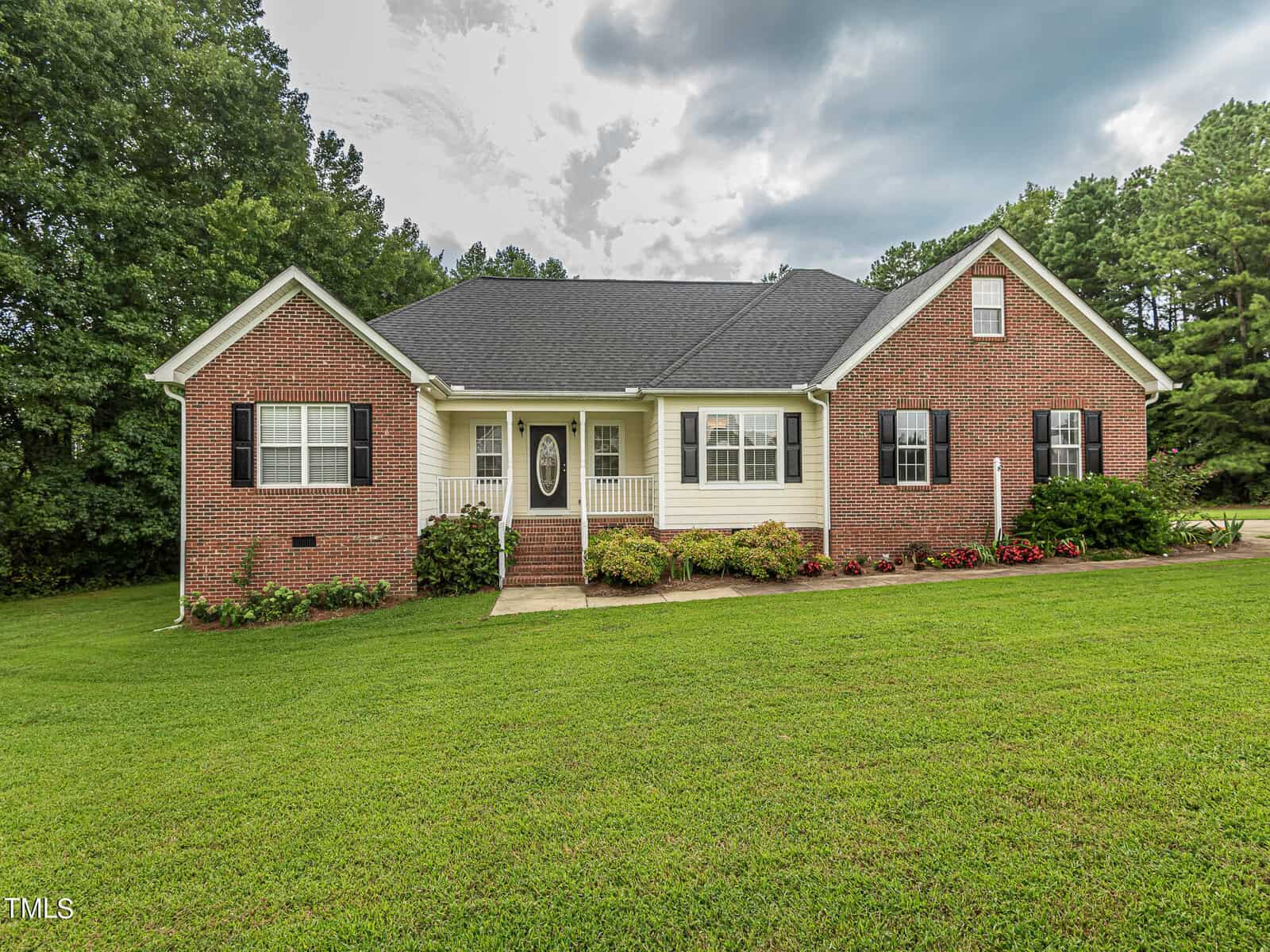 A single-story brick house at 3081 Wren Road boasts a black roof, white trim, and black shutters. The inviting front door with sidelights is framed by manicured bushes and a sprawling green lawn. Nestled in the charming community of Creedmoor, trees stand tall against a cloudy sky backdrop.