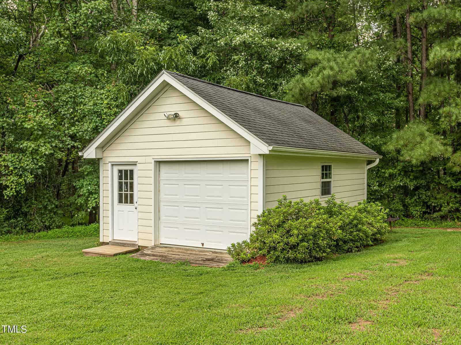 A small, single-story shed with a white garage door and side entrance stands in a lush green yard. It's surrounded by dense trees and bushes, with a grassy lawn in the foreground, under a partly cloudy sky.