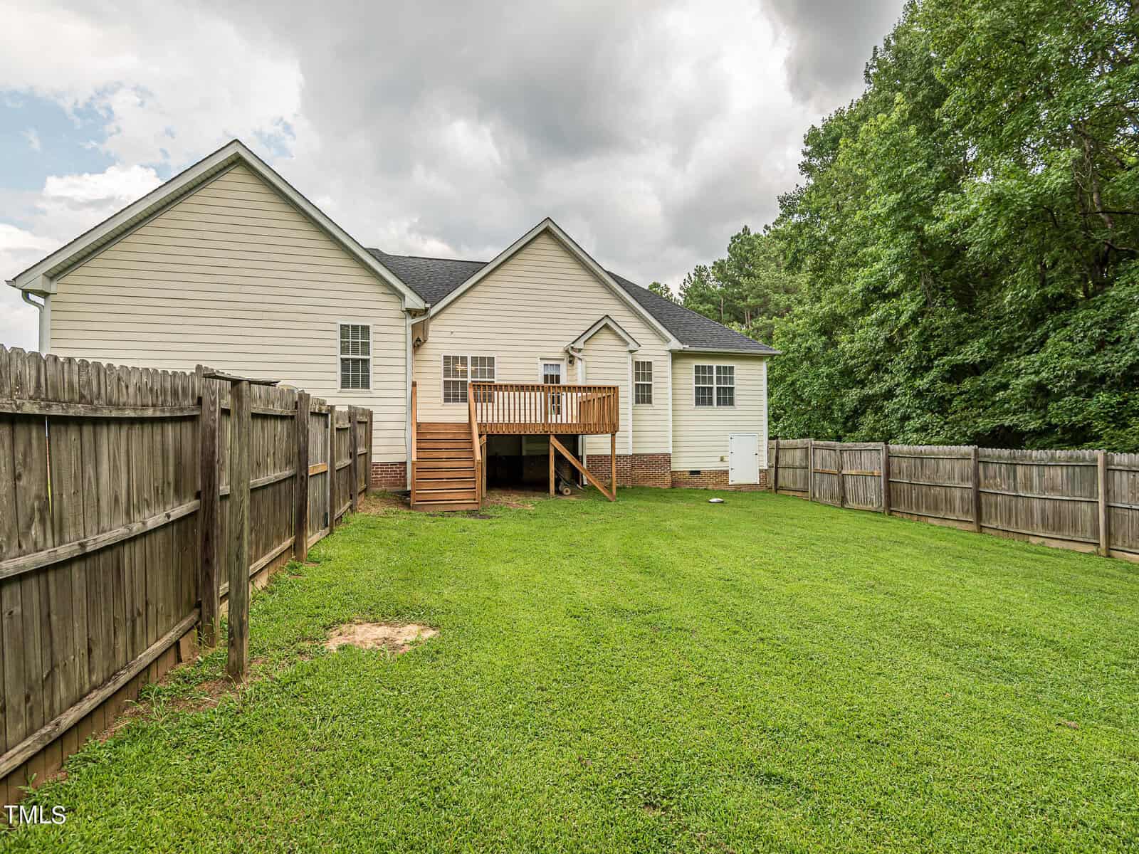 A white house with a wooden deck is surrounded by a wooden fence. The backyard features a well-kept lawn and is bordered by trees. The sky is overcast.