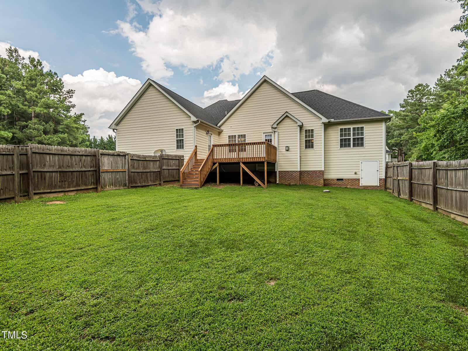 A suburban backyard with a well-maintained lawn, enclosed by a wooden fence. A two-story white house with a black roof and a small wooden deck is visible. The sky is cloudy, and trees are in the background.