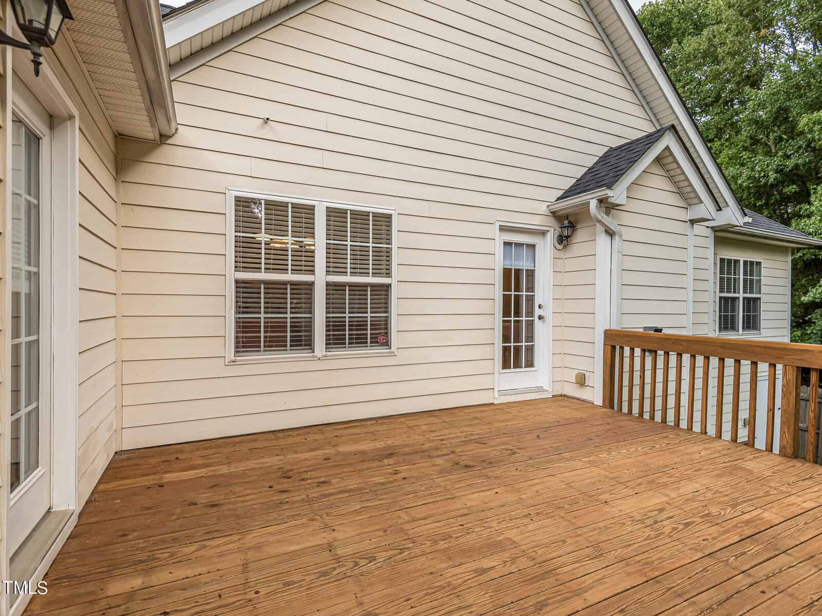 A wooden deck attached to a beige house with white-framed windows and a glass door. The deck appears spacious, surrounded by a wooden railing. The exterior has vertical siding, and trees are visible in the background.