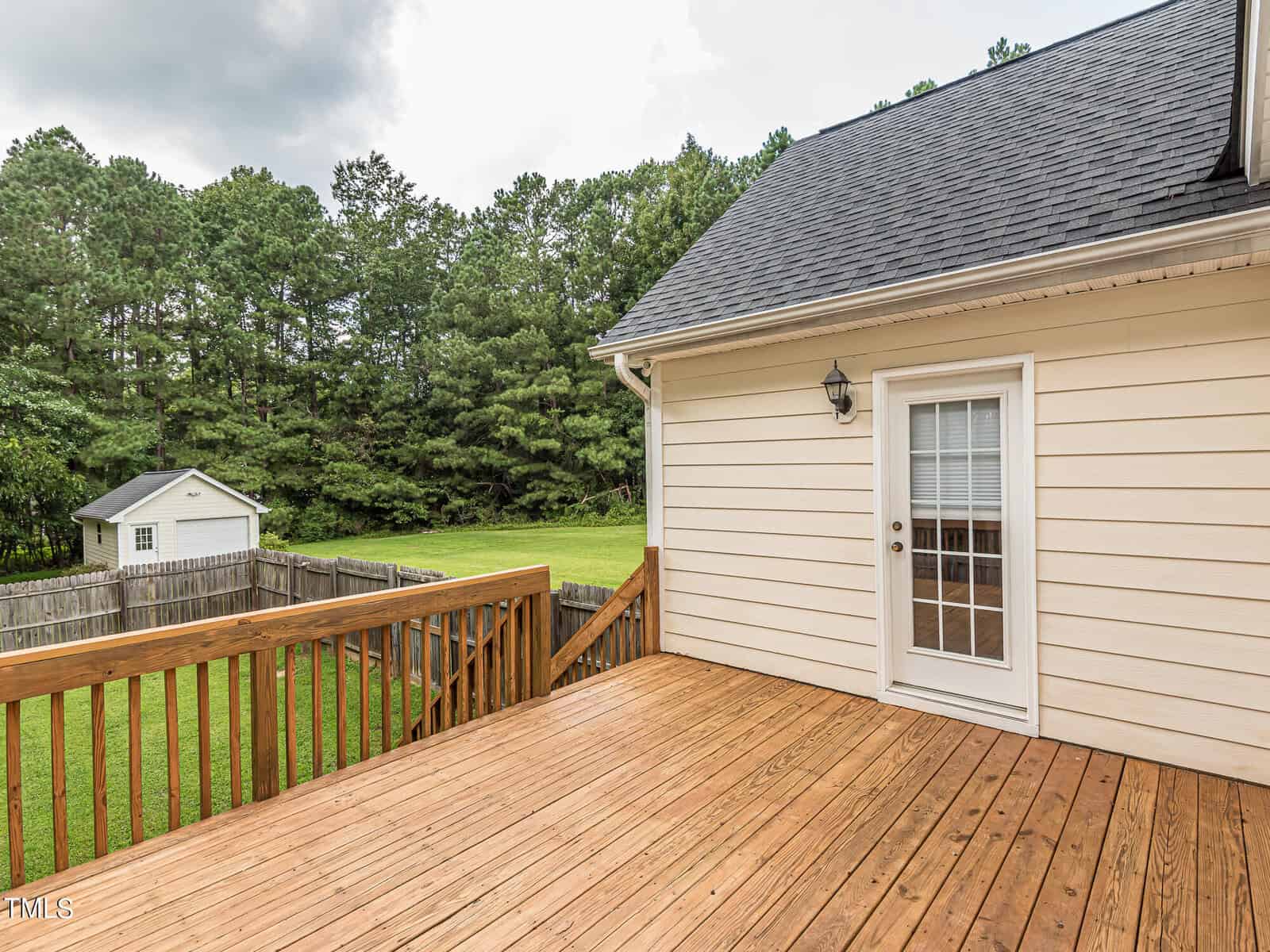 A wooden deck extends from a beige house, featuring a glass door with a light fixture above. The deck overlooks a grassy backyard surrounded by a wooden fence, with a small shed in the distance and trees lining the perimeter.