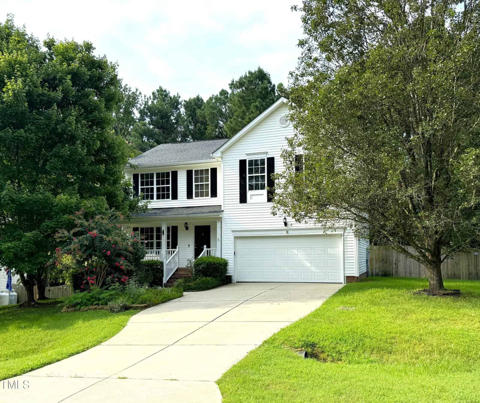 Nestled on 447 Mayview Drive, this two-story white house in Creedmoor boasts a double garage, surrounded by trees and lush greenery. The welcoming front features a quaint porch and multiple windows, with a concrete driveway leading up to the garage under a cloudy sky.