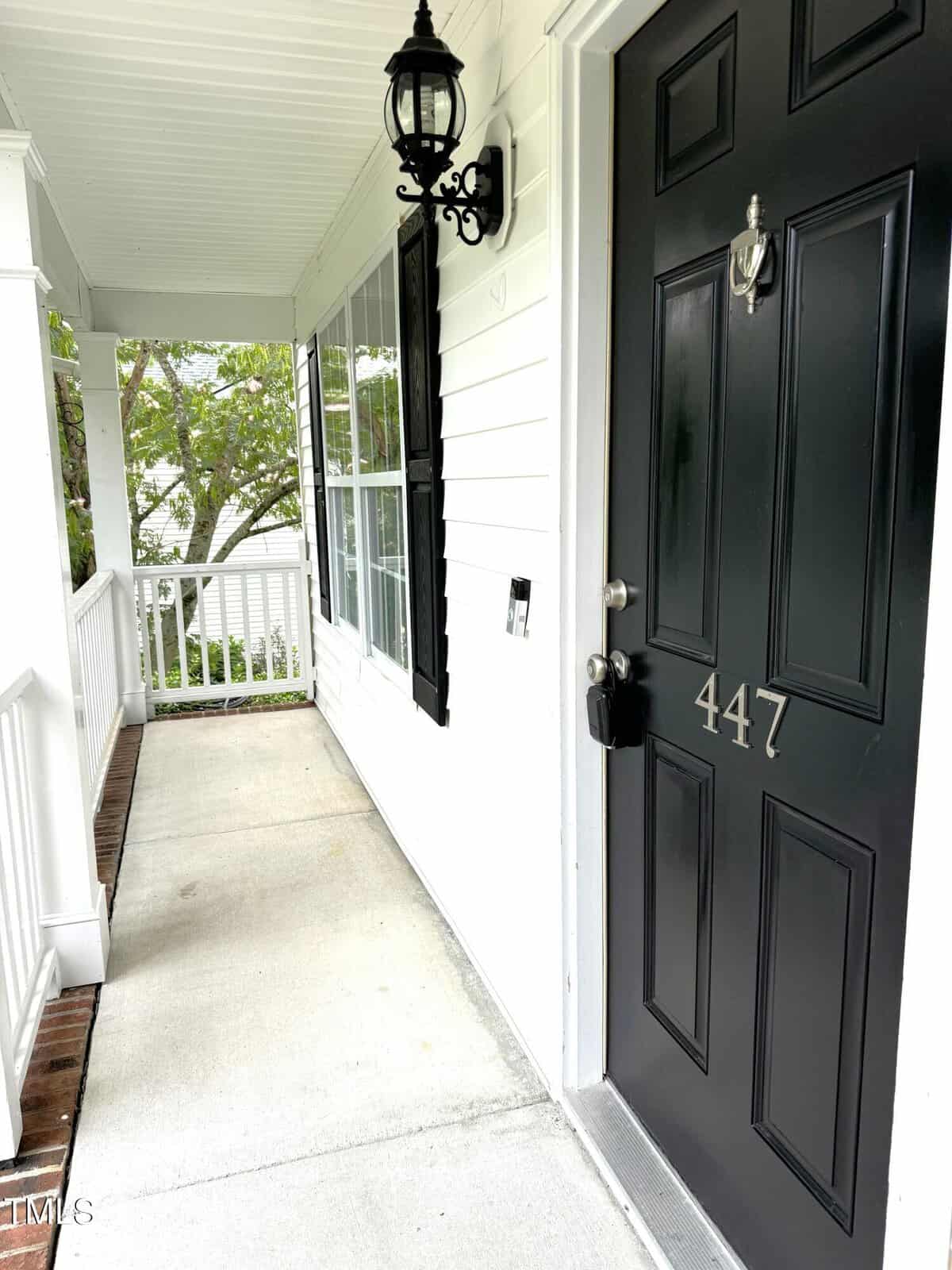 A black front door with the number 447, featuring a silver knocker and handle. The door is set in a white-paneled house with a porch, white railing, and a black lantern light fixture above. There are green trees visible in the background.