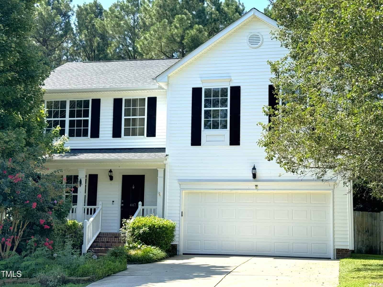 A two-story white house with black shutters, a double garage, and a front porch. There are trees and shrubs around the driveway and a well-maintained lawn in front.