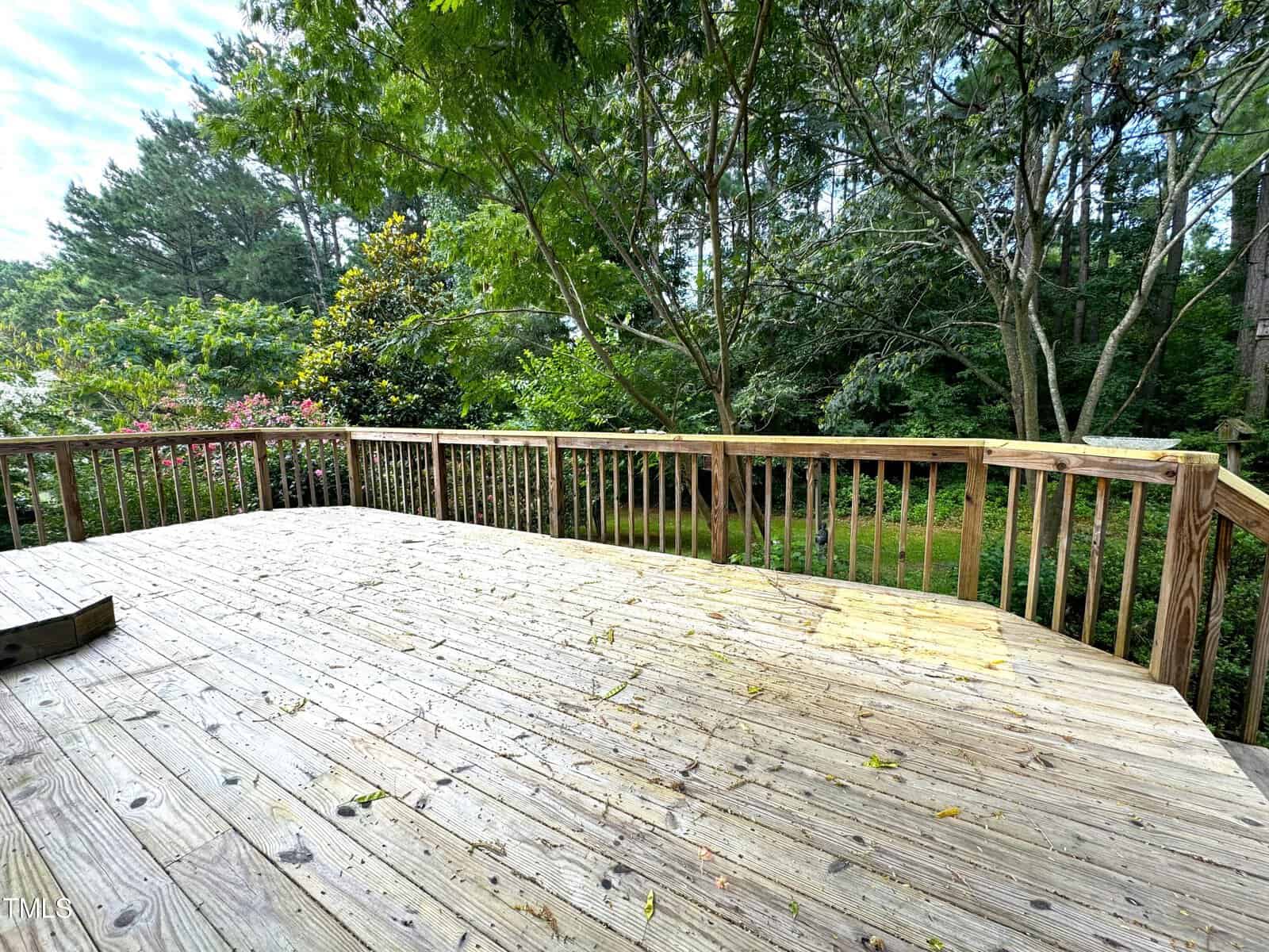 Wooden deck overlooking a lush garden with tall trees and flowering shrubs. The sky is partly cloudy, casting dappled sunlight on the scene.