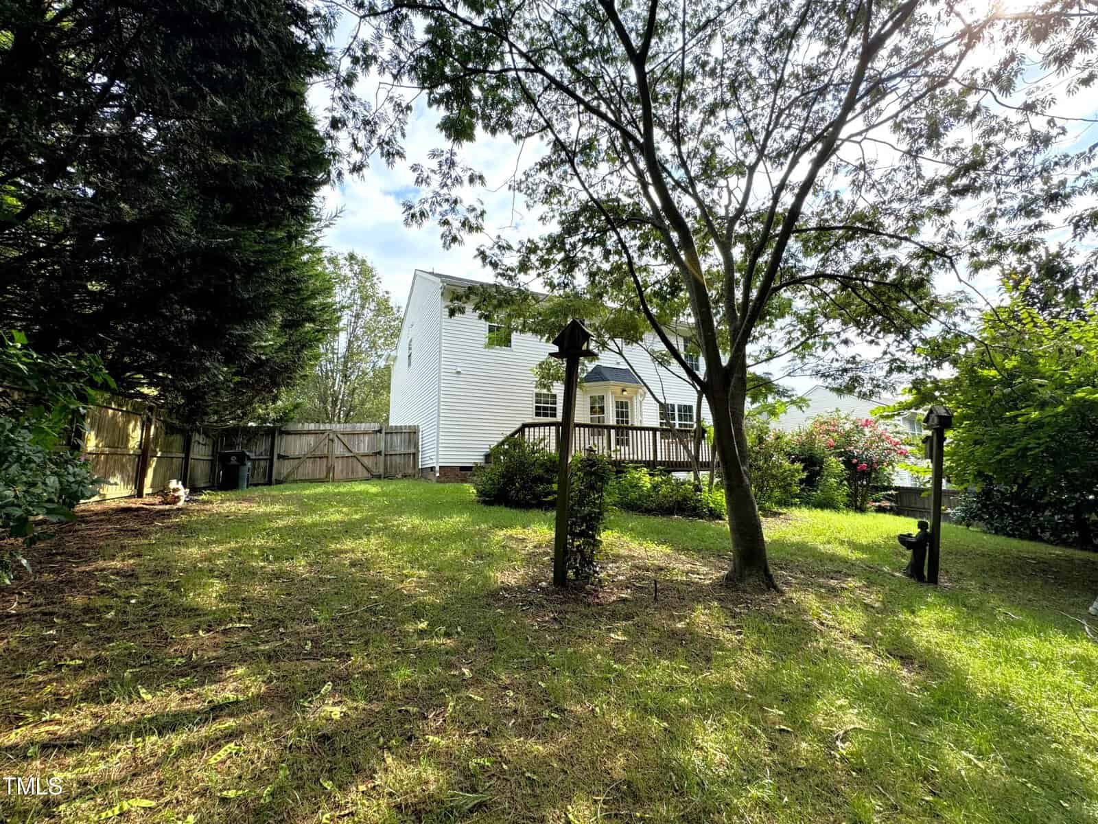 A tranquil backyard with lush green grass, trees, and a wooden birdhouse mounted on a pole. A white house with a small porch is in the background, partially obscured by foliage. A wooden fence encloses the yard.