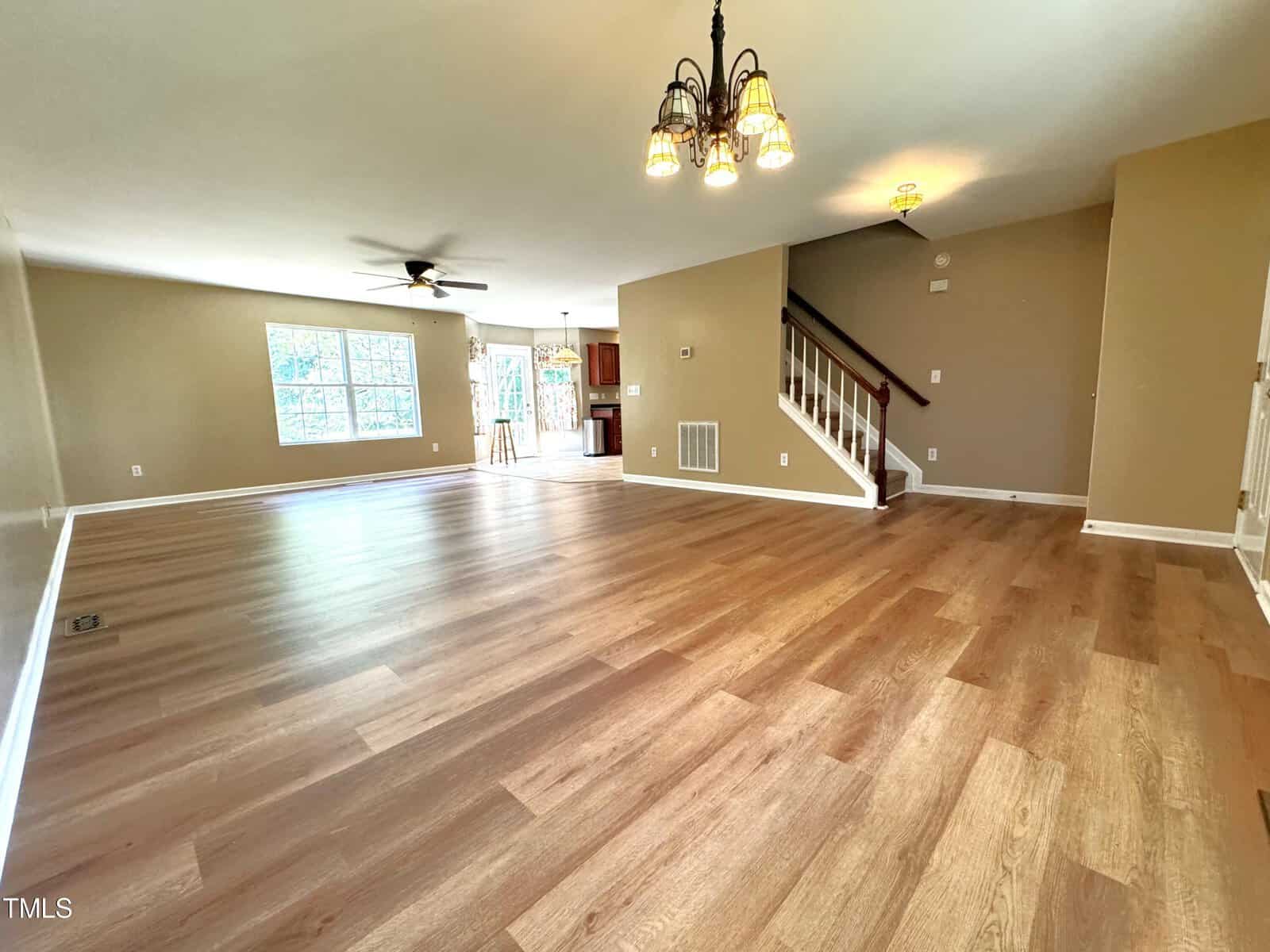 Spacious living room with light wood flooring, beige walls, and a ceiling fan. A black chandelier hangs above the open area, while wide windows let in natural light. A staircase leads to the upper floor.