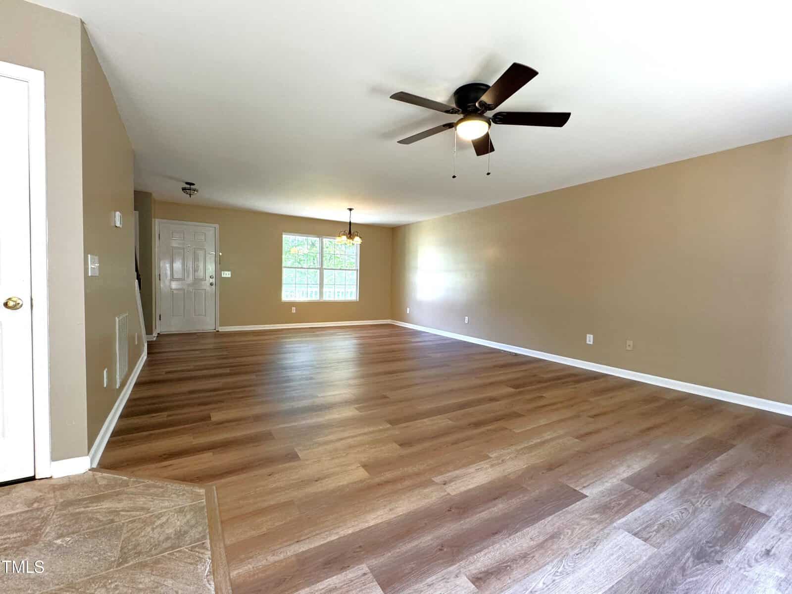 A spacious, empty living room with light wood flooring, beige walls, and a ceiling fan. A large window and a small hanging light fixture brighten the room. The entrance door is visible in the background.