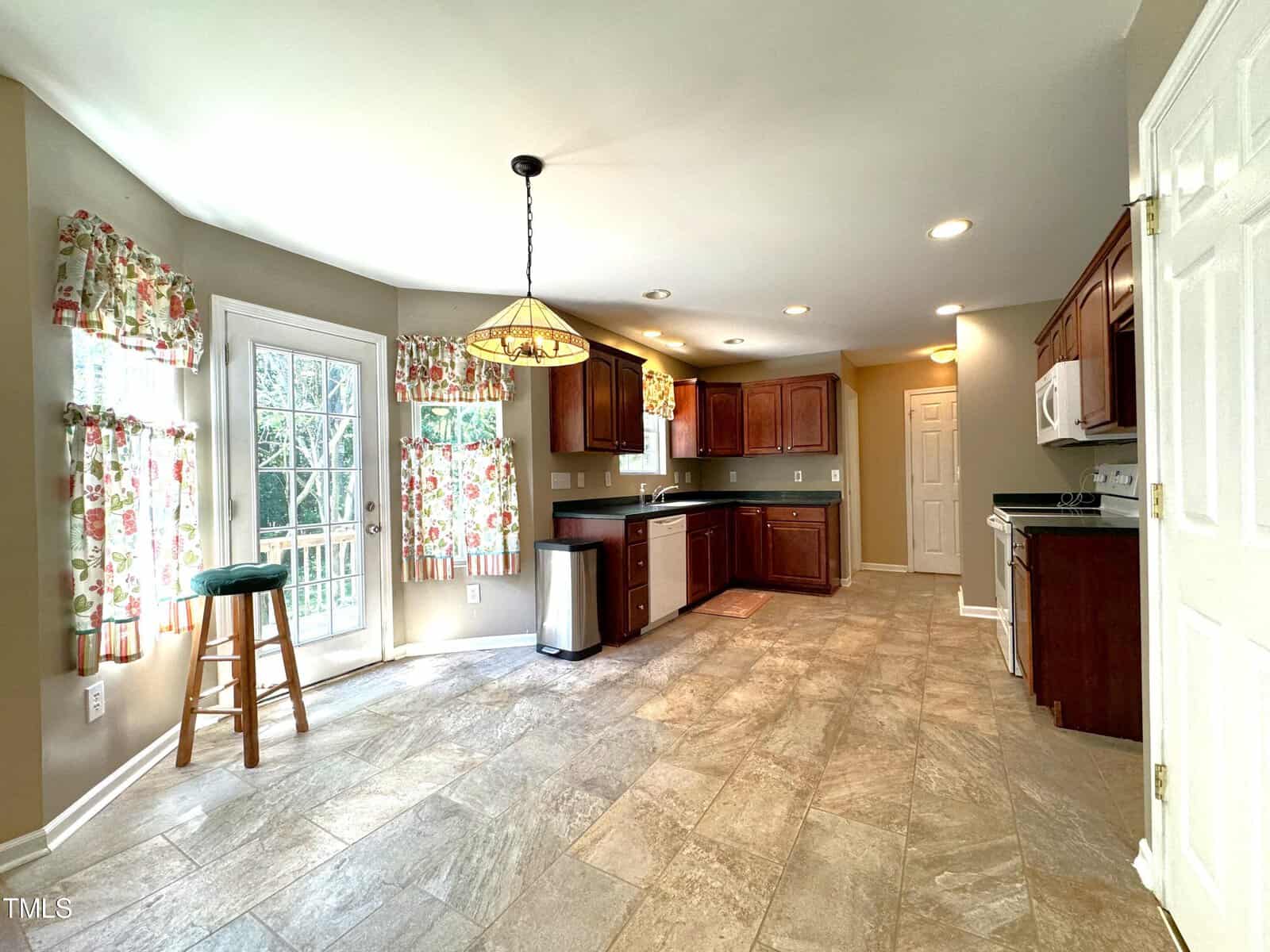 Bright kitchen with tiled floors, wooden cabinets, and floral curtains. Features a central light fixture, a stool by the windowed door, and stainless steel appliances. Ample natural light fills the room.