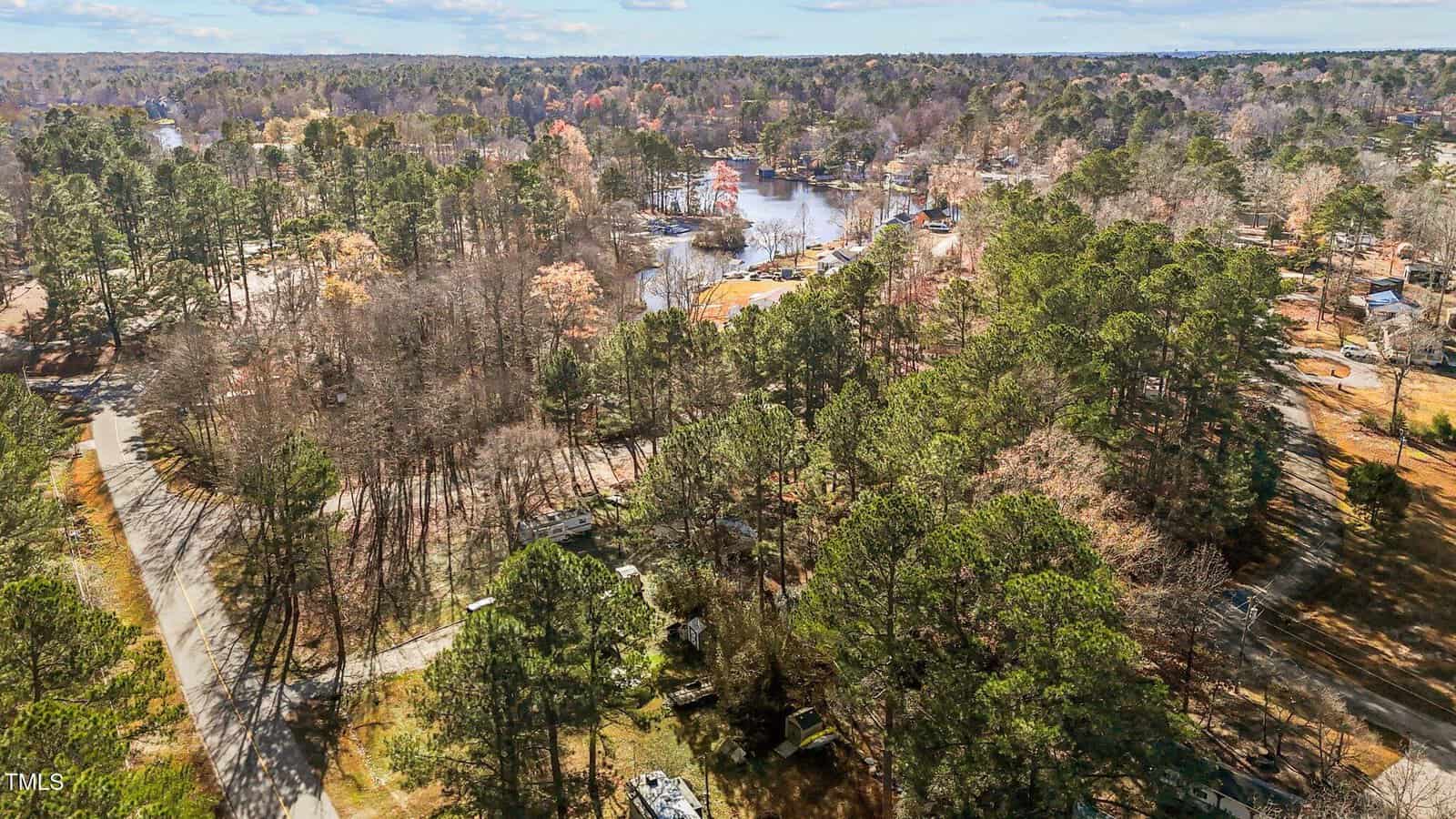 Aerial view of a wooded neighborhood with scattered houses, roads, and a nearby lake. Trees dominate the landscape, and the water is visible in the background, reflecting the sky.