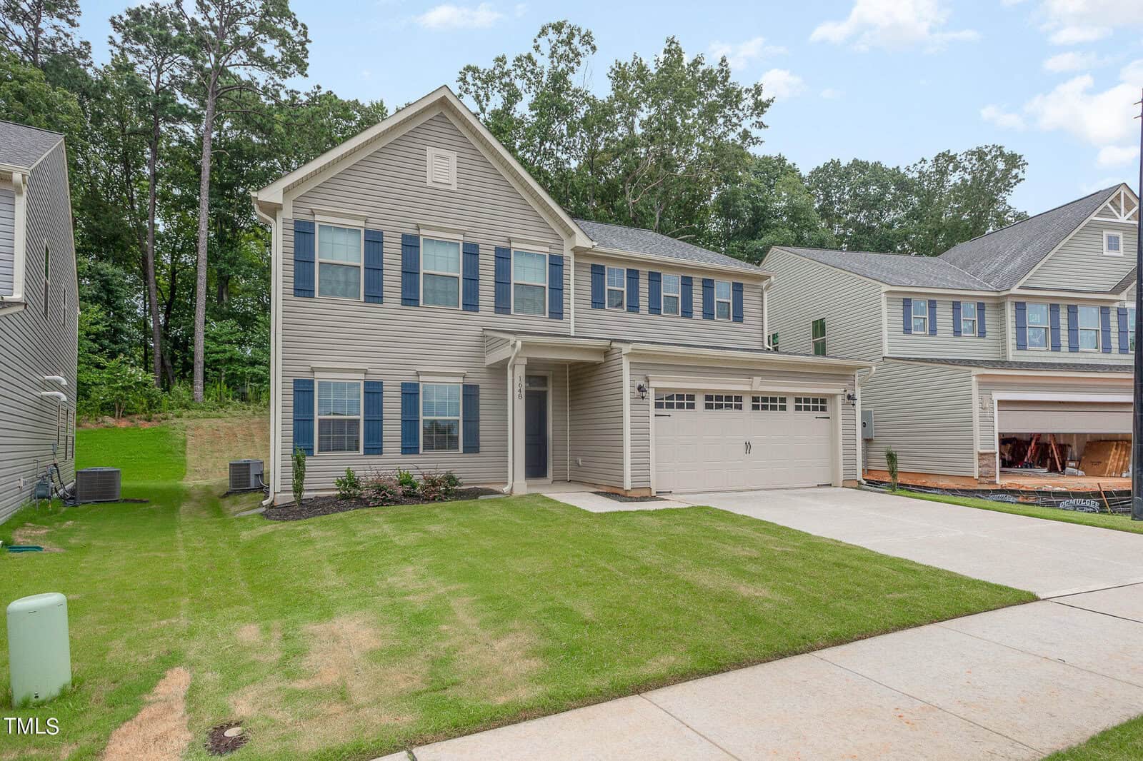 Two-story beige suburban house with blue shutters, a double garage, and a neatly mowed lawn. Neighboring houses and trees are visible in the background under a partly cloudy sky.