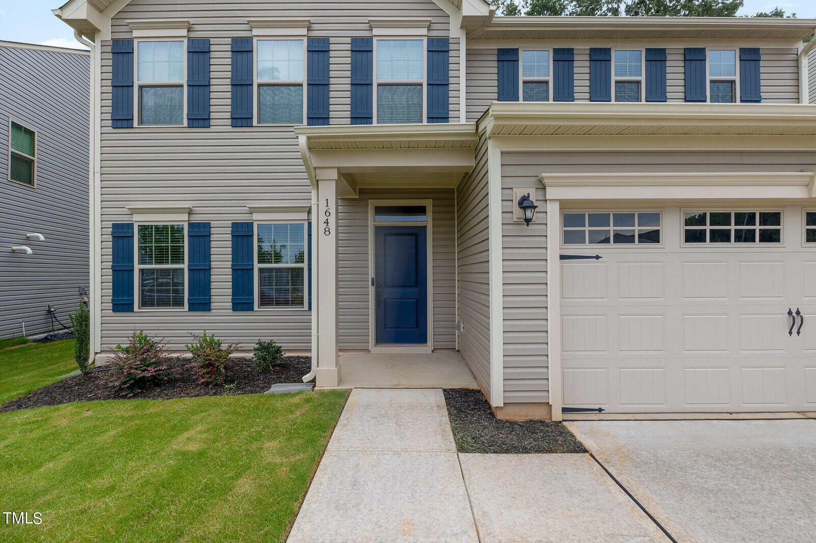 Front exterior of a beige two-story house with blue shutters, a blue front door, attached garage, manicured lawn, and small shrubs by the entrance.